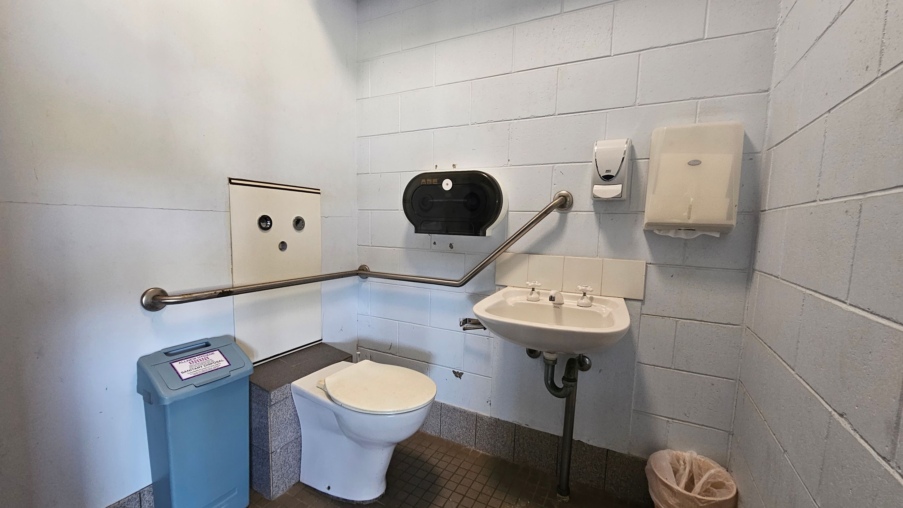 A public toilet with light grey tiled walls and a small square tiled floor. On the left, there's a blue sanitary disposal bin and a white toilet with a metal grab rail. Above the toilet is a black toilet paper dispenser. On the right, a white sink with two taps is mounted on the wall, accompanied by a soap dispenser and a paper towel holder. A small waste bin is on the floor beneath the sink.