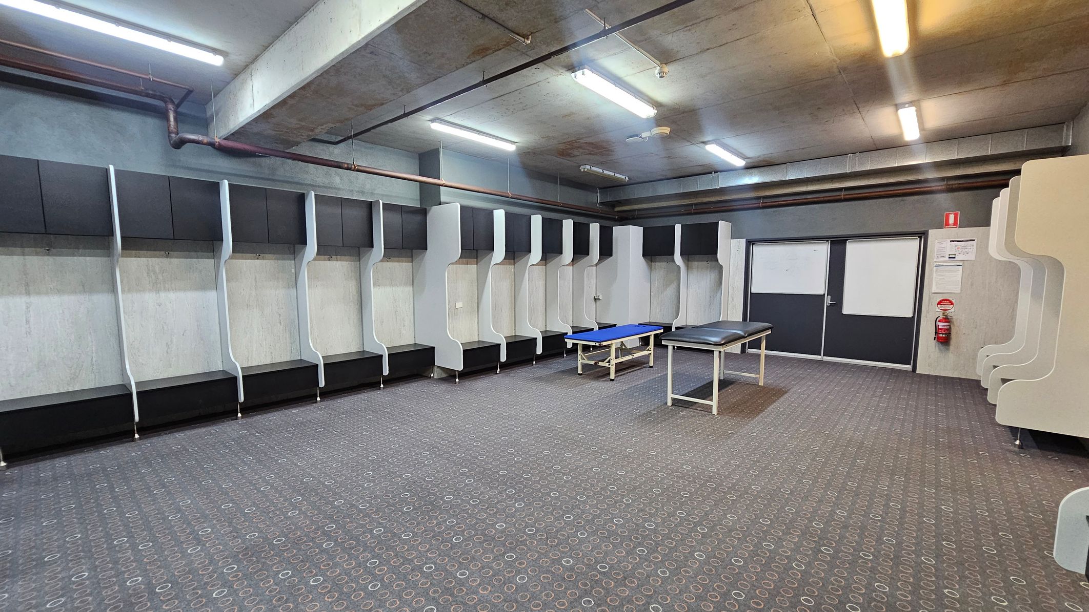A spacious locker room with a patterned carpeted floor. On the left and centre, there are multiple open lockers with black and white sections. In the middle, two examination tables are placed on the floor. The right side features a set of double doors with a fire extinguisher and notices on the wall. The ceiling is concrete with exposed pipes and fluorescent lighting.