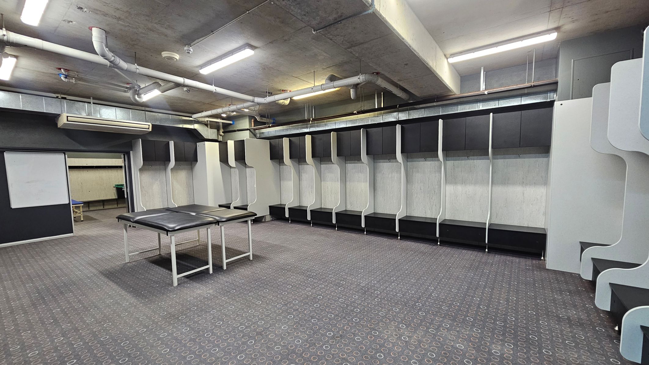 A spacious locker room with a patterned carpeted floor. On the right, there are several open lockers with black and white panels. In the centre, two massage tables with black padding are positioned. The ceiling is industrial, featuring exposed pipes and fluorescent lighting. In the background, a doorway leads to another room with a table visible. The walls are a mix of dark and light colours, and an air conditioning unit is mounted on the wall.