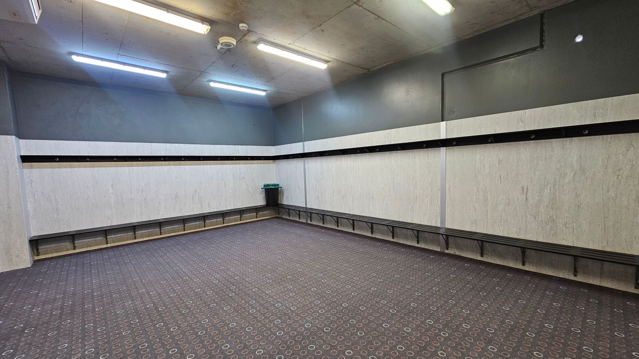 A room with grey walls and a patterned carpet floor featuring circular designs. Along the walls, there are black benches with hooks above them. In the corner, a green bin is placed. The ceiling is grey with fluorescent lights and a smoke detector.
