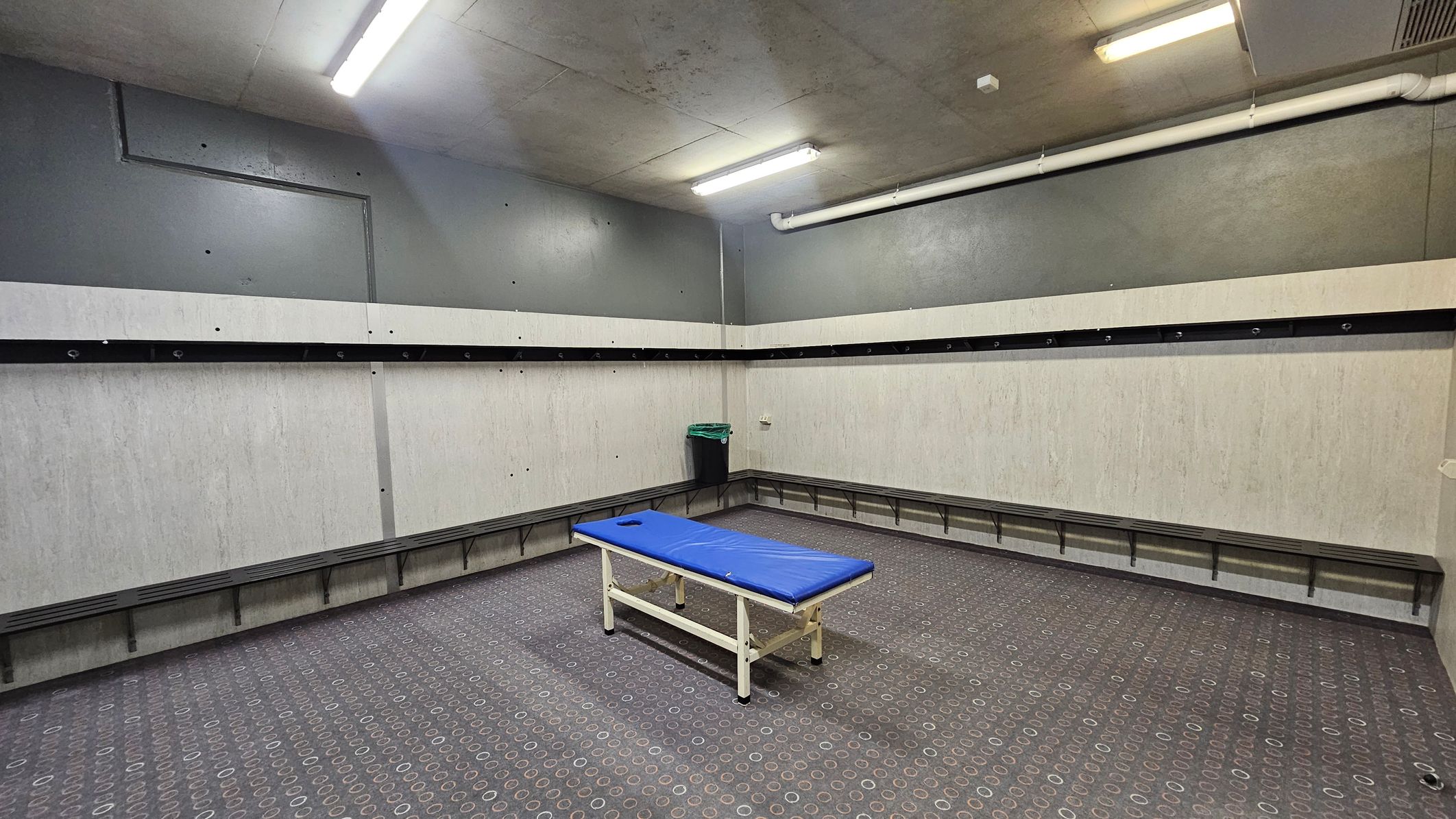 A room with grey walls and a patterned carpeted floor. In the centre, there's a blue padded bench or massage table. Along the walls, there are long benches with hooks above them, likely for hanging items. A small green bin is in the corner. The ceiling has fluorescent lights and a visible white pipe running along it.