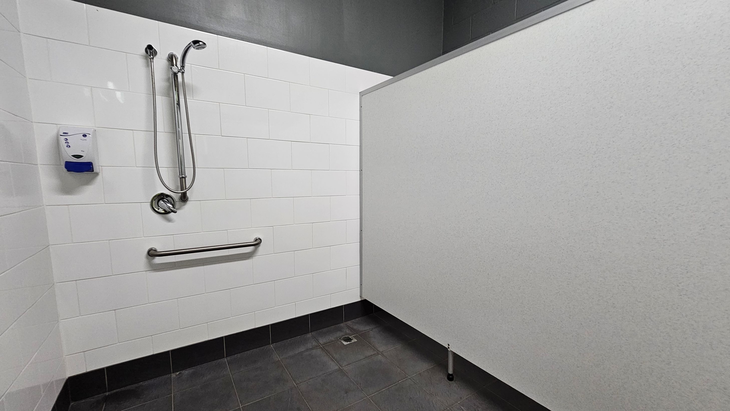 A shower area with white tiled walls on the left, featuring a mounted showerhead and a handheld shower attachment. Below the shower is a horizontal metal grab bar. A soap dispenser is mounted on the wall. The floor is dark grey with a central drain. On the right, there is a light grey partition wall. The ceiling is dark grey.