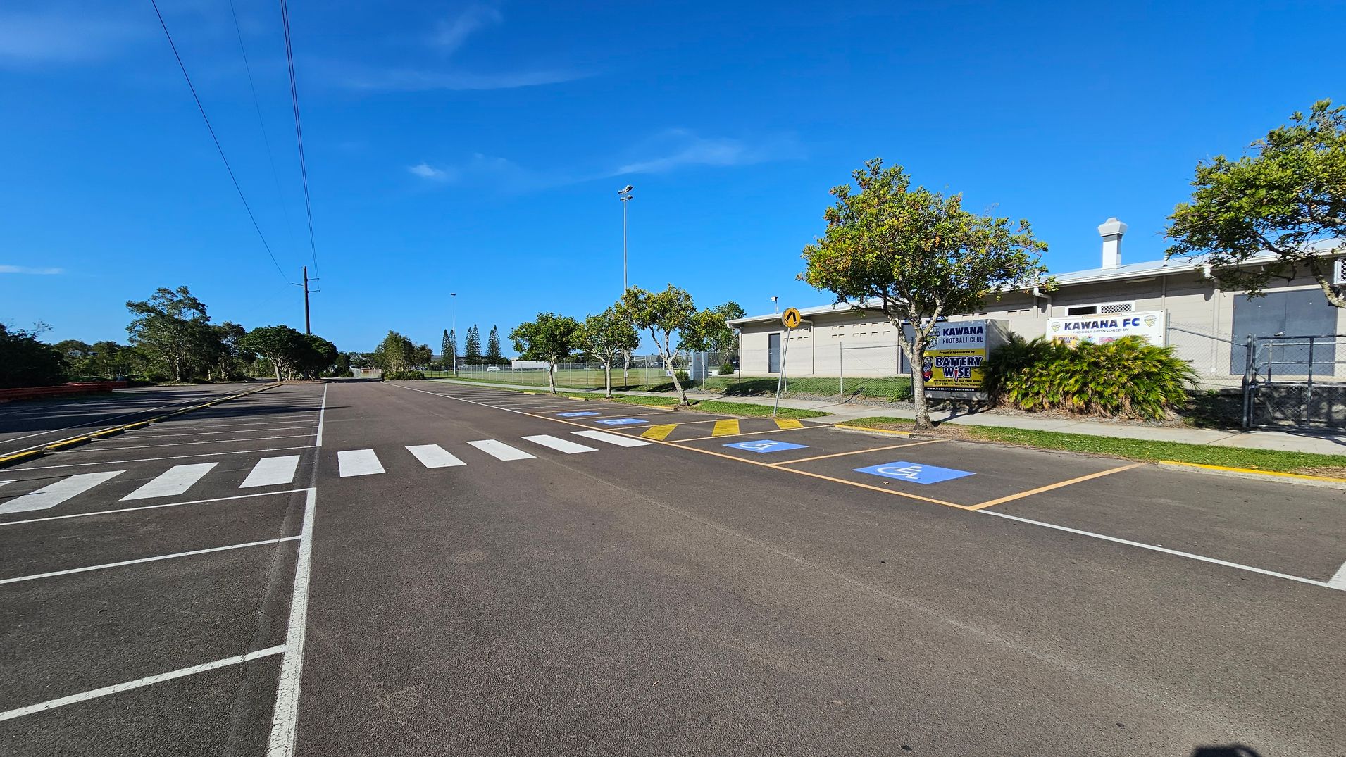 A parking area with marked spaces, including disabled parking, and a zebra crossing in the foreground. On the right, a building with signs for "Kawana Football Club" and "Kawana FC" is partially obscured by trees and shrubs. Several small trees line the pavement. In the background, a fence and more trees are visible. The sky is clear and blue.