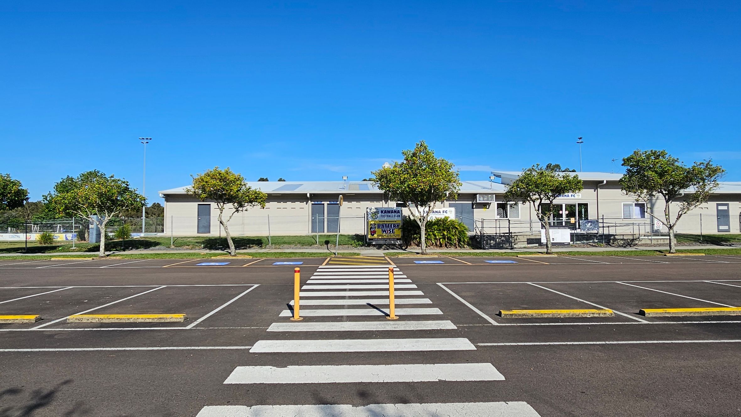 A parking area with marked spaces and a zebra crossing in the foreground. Two yellow bollards are positioned at the crossing. In the background, a single-storey building with a light-coloured facade is visible, flanked by several small trees. Signs on the building read "Kawana Football" and "Kawana FC". The sky is clear and blue.