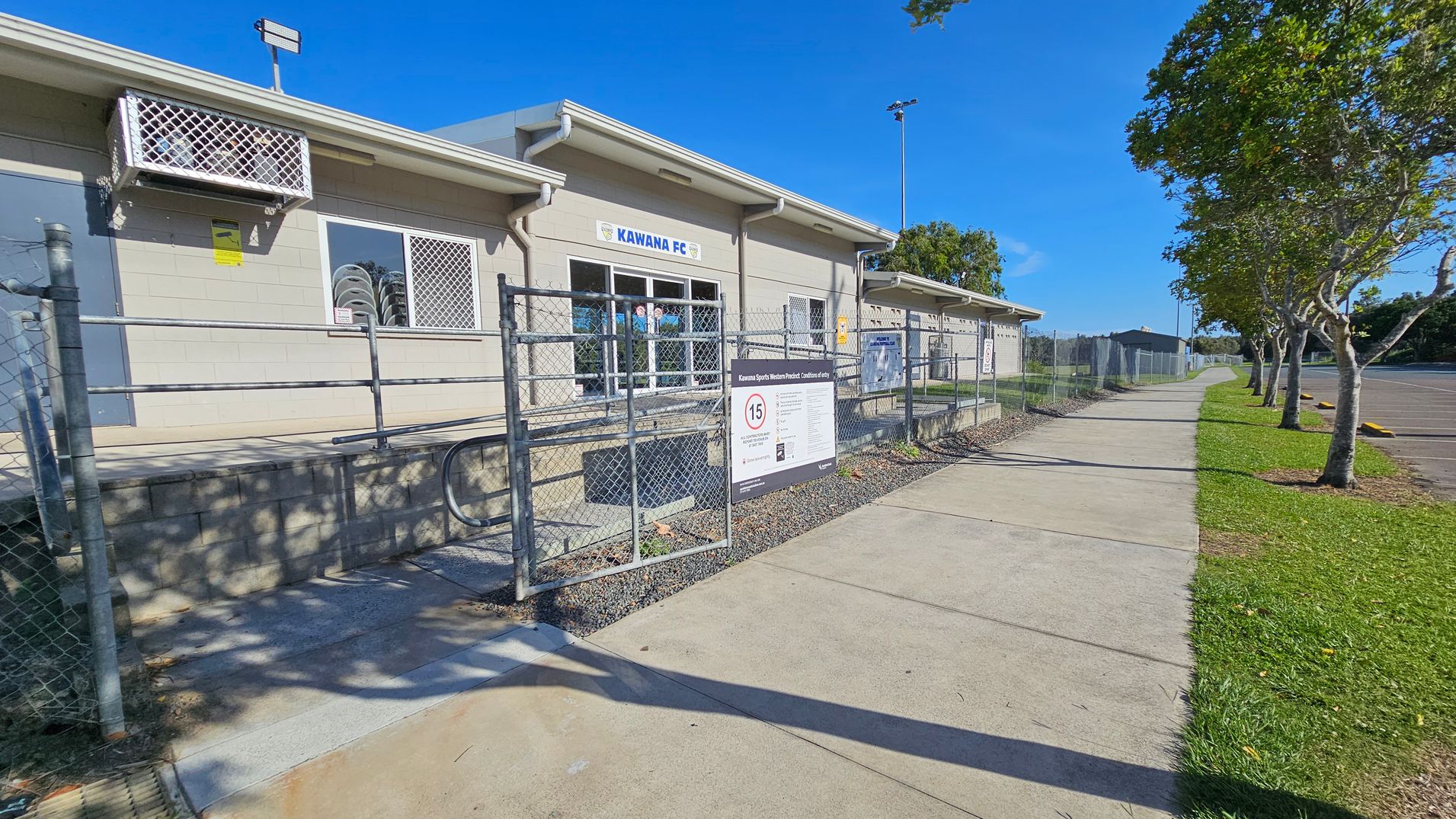 A single-storey building with a sign reading "Kawana FC" is on the left. It has barred windows and an air conditioning unit. A chain-link fence runs along the front, with a sign about entry conditions. A concrete path leads to the right, bordered by grass and a row of trees. The sky is clear, and there is a car park visible in the background on the right.