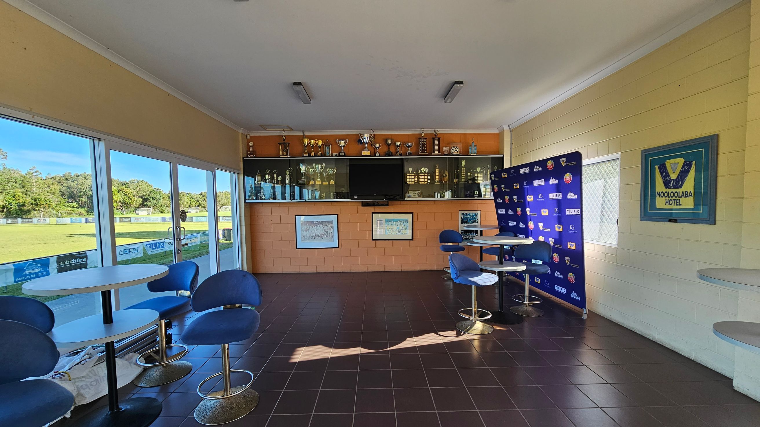 A room with large glass windows on the left, offering a view of a grassy field. Inside, there are round tables with blue chairs. The back wall features a display of trophies and a large TV. A framed sports jersey with "Mooloolaba Hotel" is on the right wall, alongside a blue backdrop with logos. The floor is tiled in dark brown, and the ceiling is white with fluorescent lights.