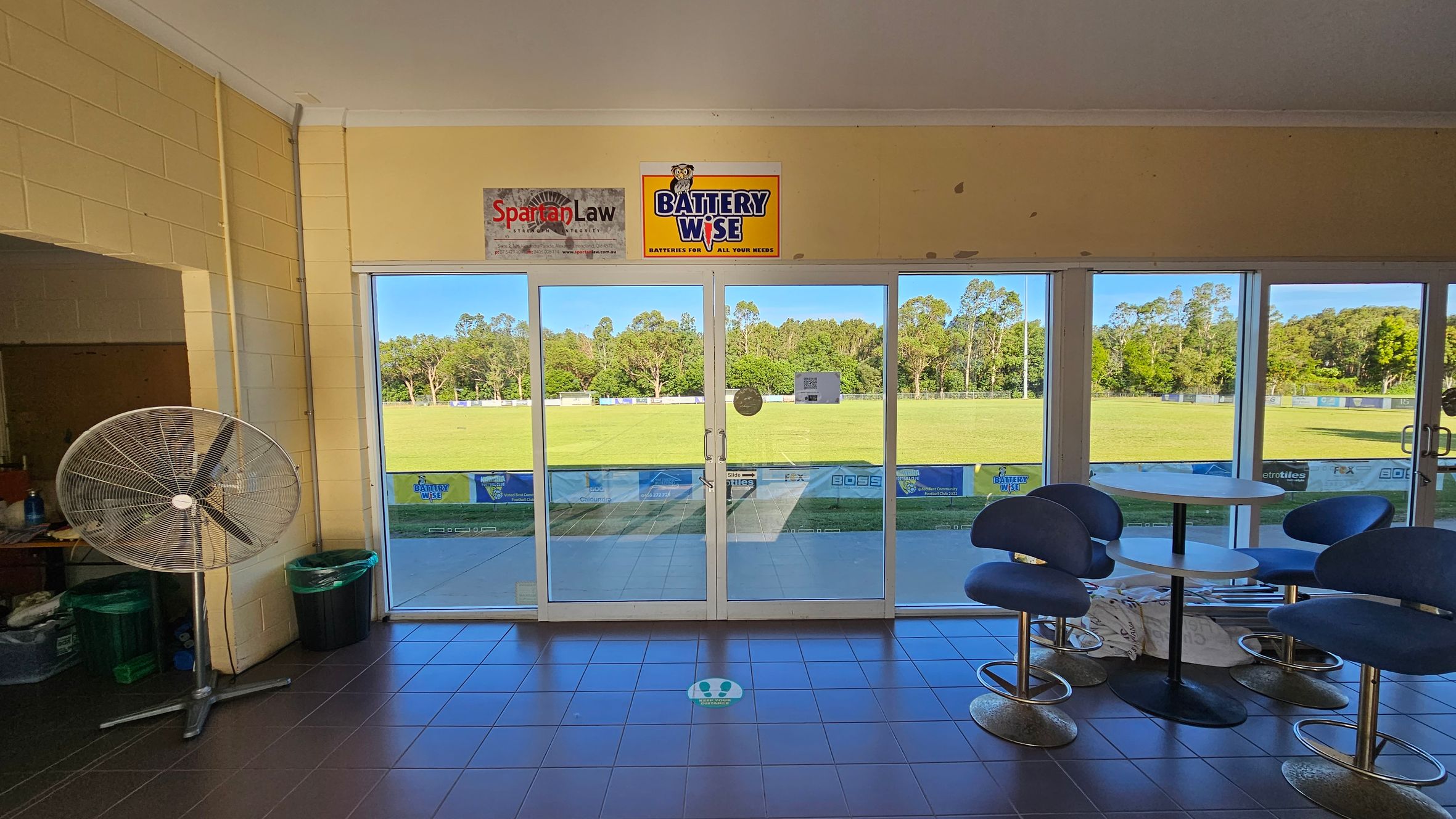 A room with large glass doors in the centre, leading to a grassy field outside. On the left, there's a large standing fan and a couple of bins. On the right, there are several blue chairs around a round table. The floor is tiled in dark grey, and there are two signs above the doors, one for "Spartan Law" and another for "Battery Wise". Trees are visible in the background through the glass doors.