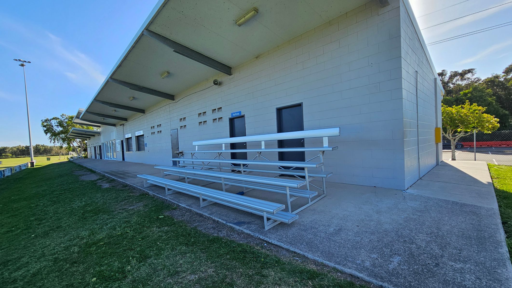 A sports facility with a long, light-coloured building on the right. The building has several doors and small windows, with a sign marked "referee" above one door. In front of the building are metal bleachers on a concrete surface. To the left, there is a grassy area with a tall floodlight and trees in the background. The roof of the building extends outward, providing some shade. A small tree and a fence are visible in the background on the right.