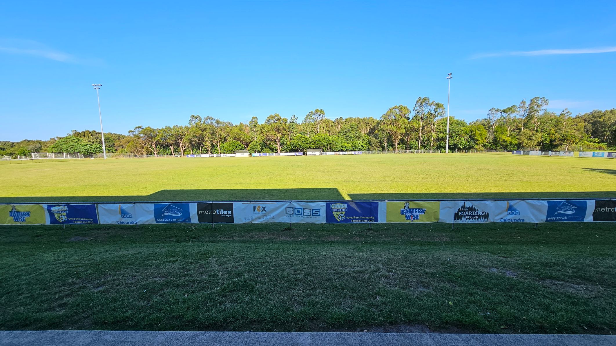 A grassy sports field with a row of advertising banners in the foreground. The field is expansive, with two tall floodlights on either side. In the background, there are trees lining the perimeter, providing a natural backdrop. The sky is clear, and the field is well-maintained.