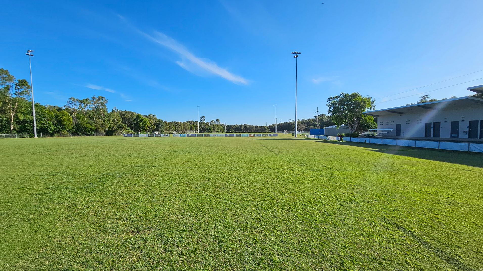 A grassy sports field with tall floodlights on the left and right. In the background, there are trees lining the perimeter. On the right, a building with a flat roof and several doors is visible, along with a few trees nearby. The field is expansive and well-maintained.