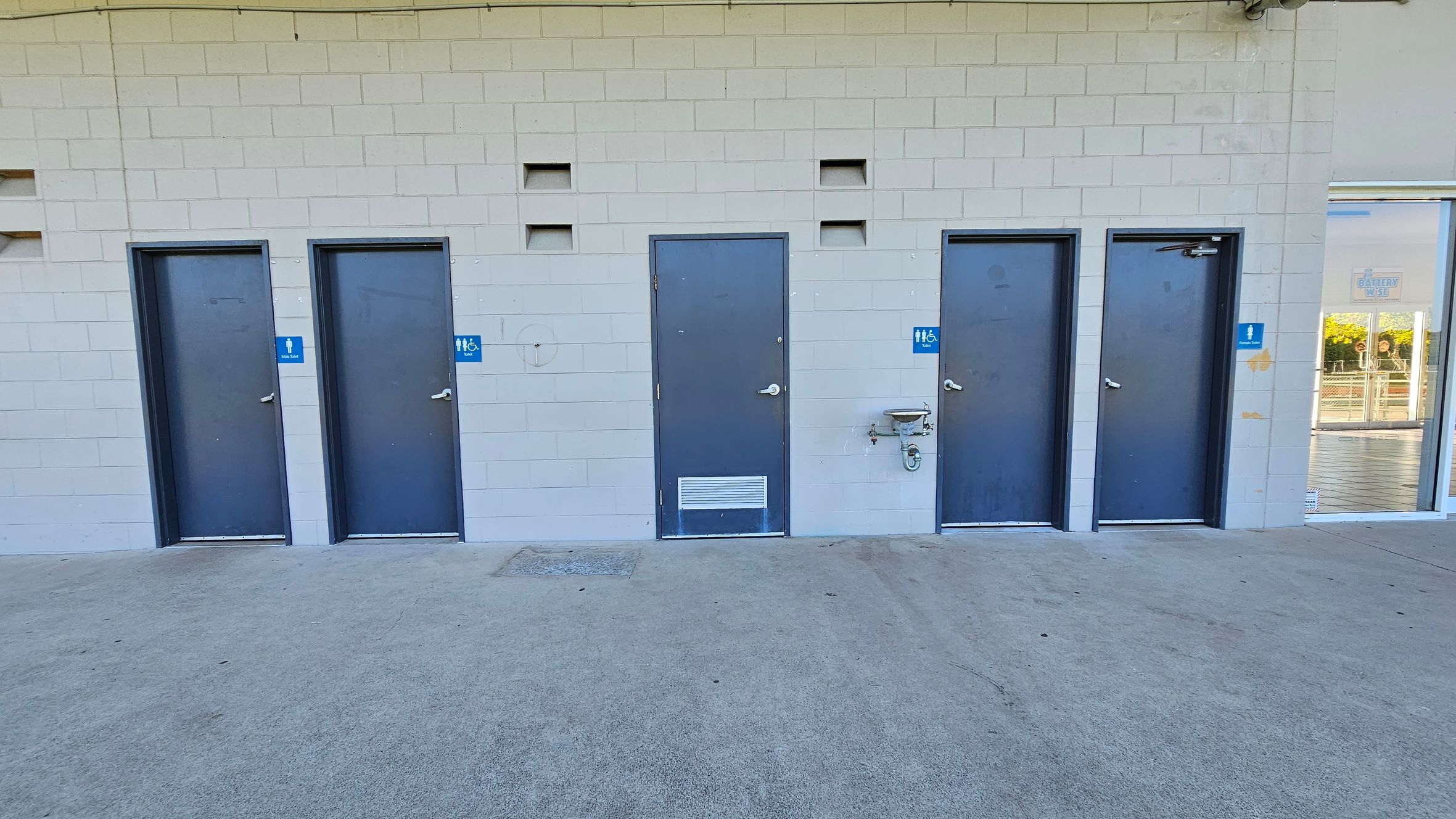 A row of four grey doors set in a light-coloured brick wall, each with a blue sign indicating a toilet. The second and third doors from the left have additional signs for accessibility. Between the third and fourth doors, there is a drinking fountain attached to the wall. On the far right, a glass door reveals a glimpse of an interior space with a sign that reads "BATHING AREA." The ground is concrete, and the ceiling is not visible.