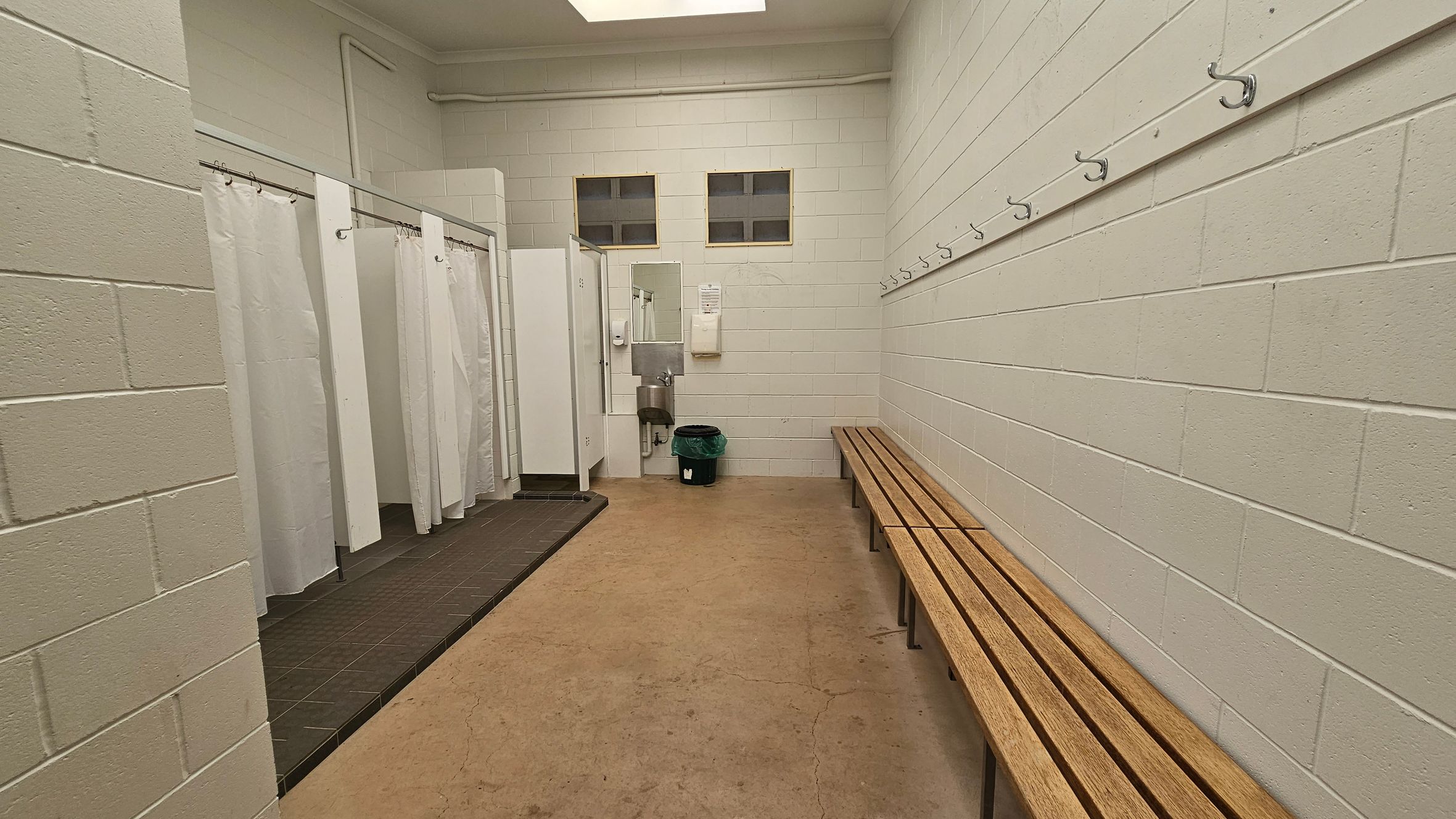 A changing room with white brick walls. On the left, there are several shower stalls with white curtains. In the middle, a sink with a mirror above it and a waste bin below. On the right, a long wooden bench runs along the wall, with hooks above it. The floor is concrete, and the ceiling is plain with a light fixture.