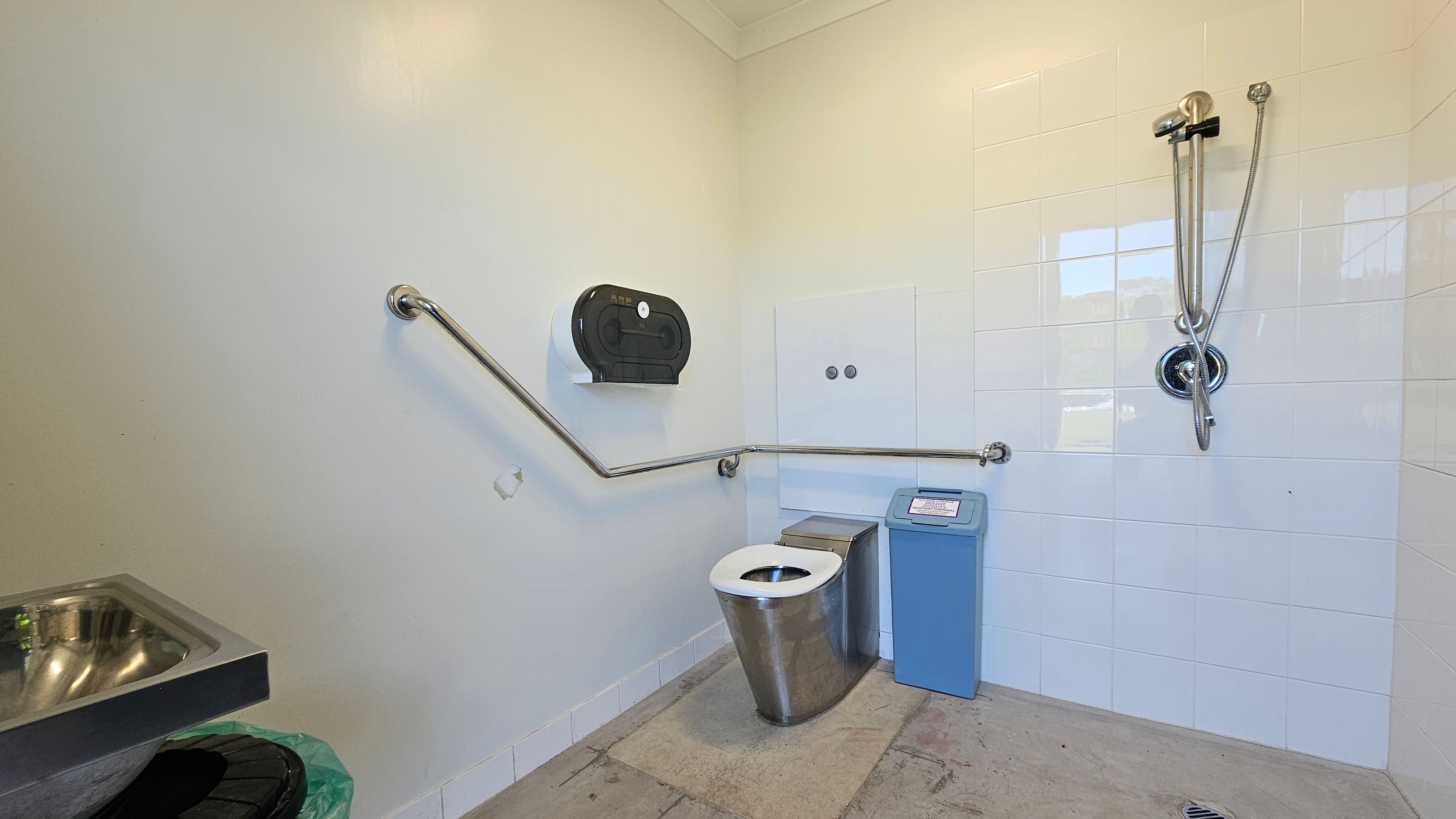 A public restroom with a stainless steel toilet in the centre, equipped with a blue sanitary bin to the right. The walls are partially tiled in white, with a shower fixture on the right wall. A horizontal grab rail runs along the walls, and a black toilet paper dispenser is mounted above the toilet. On the left, a stainless steel sink is visible, with a green bin beneath it. The floor is concrete, and the ceiling is plain.