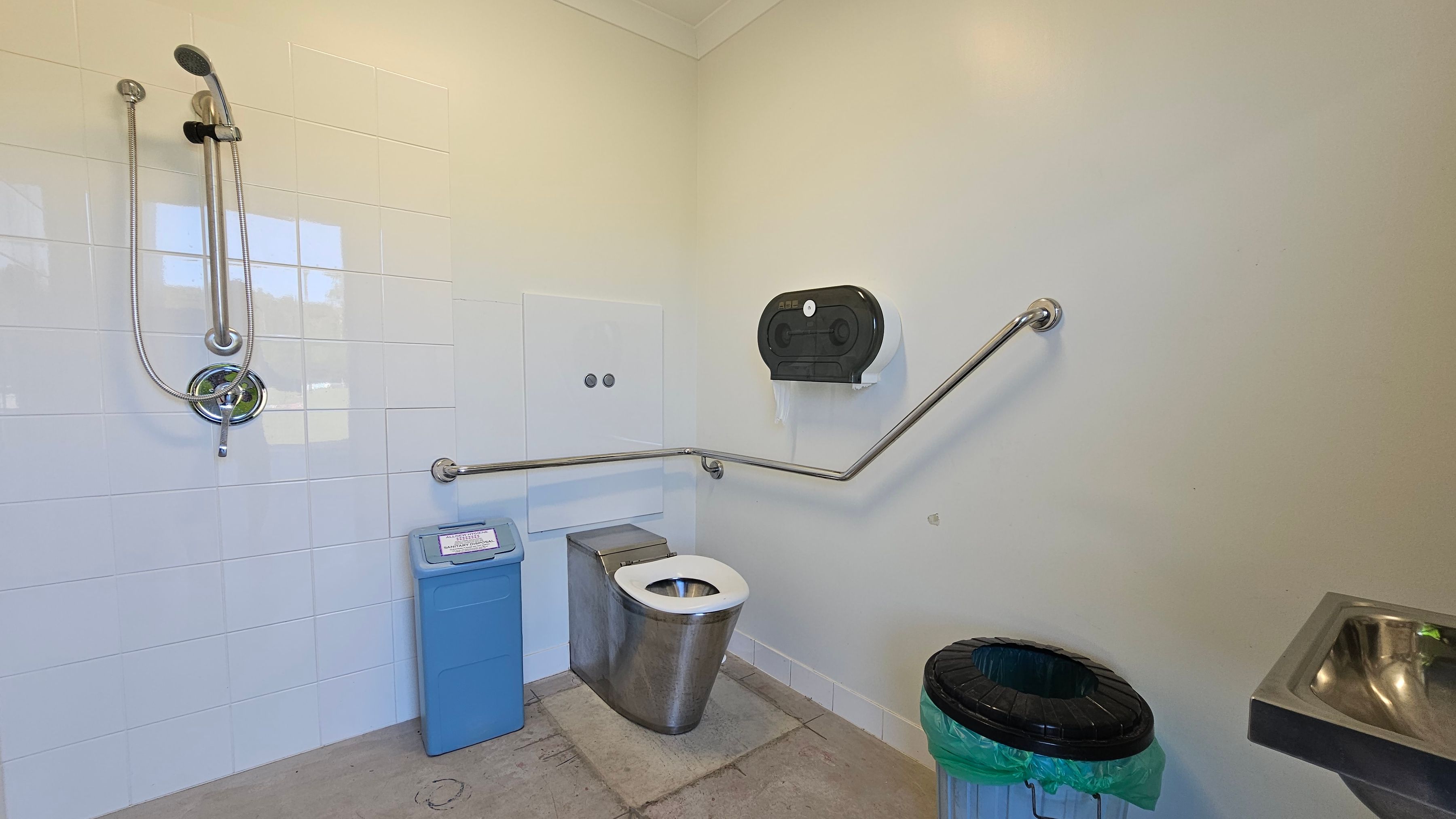 A public restroom with a stainless steel toilet in the centre, equipped with a wall-mounted grab bar for accessibility. To the left, a showerhead is mounted on white tiled walls, with a blue sanitary bin below. On the right, a black toilet paper dispenser is mounted on the wall, and a green-lined rubbish bin is on the floor. A small stainless steel sink is partially visible on the far right. The floor is concrete, and the walls are painted a light colour.