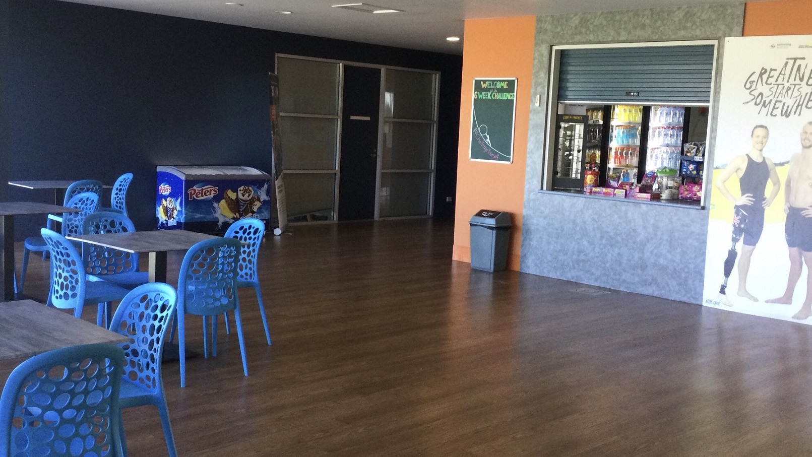 The centre’s café offers a limited selection of drinks, chips, candies, and other snacks. A large poster with images of both male and female fitness models and the phrase "Greatness starts somewhere" at the top is located on the right side of the kiosk. The other side of the food stall is a green board that has a sketch with a title “Welcome to the Six-Week Challenge”. Below it is a trash bin. The seating area has blue chairs and grey square tables that can be both moved around the café area. The centre’s café offers a limited selection of drinks, chips, candies, and other snacks. A large poster with images of both male and female fitness models and the phrase "Greatness starts somewhere" at the top is located on the right side of the kiosk. The other side of the food stall is a green board that has a sketch with a title “Welcome to the Six-Week Challenge”. Below it is a trash bin. The seating area has blue chairs and grey square tables that can be both moved around the café area.