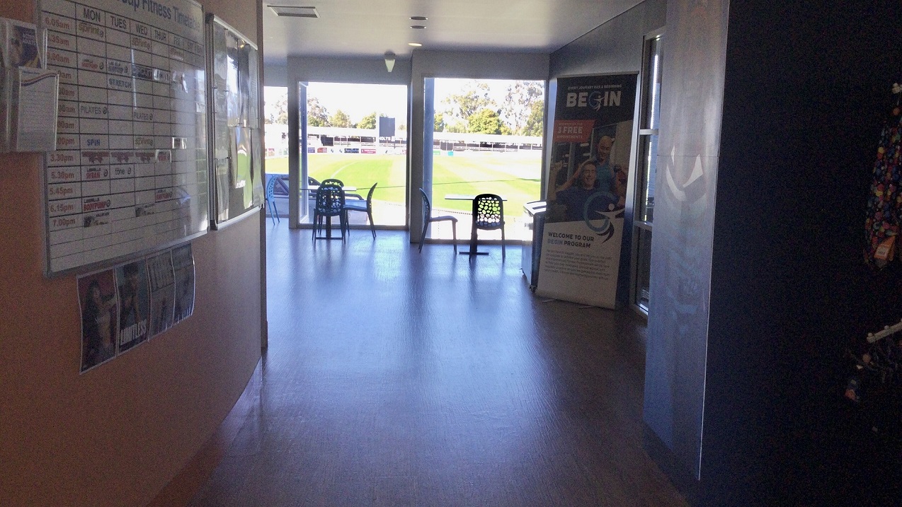 This is the extended section of the leisure centre entrance that highlights the orange wall that has the Fitness Timetable posted on the large white board. There are also flyer racks mounted on the wall where flyers are displayed. There are sets of tables and chairs placed near the glass walls where you will enjoy a pleasant green scenery. A standee of a human-sized poster is placed in the middle of this section where the word “BEGIN” is highlighted. This is the extended section of the leisure centre entrance that highlights the orange wall that has the Fitness Timetable posted on the large white board. There are also flyer racks mounted on the wall where flyers are displayed. There are sets of tables and chairs placed near the glass walls where you will enjoy a pleasant green scenery. A standee of a human-sized poster is placed in the middle of this section where the word “BEGIN” is highlighted.