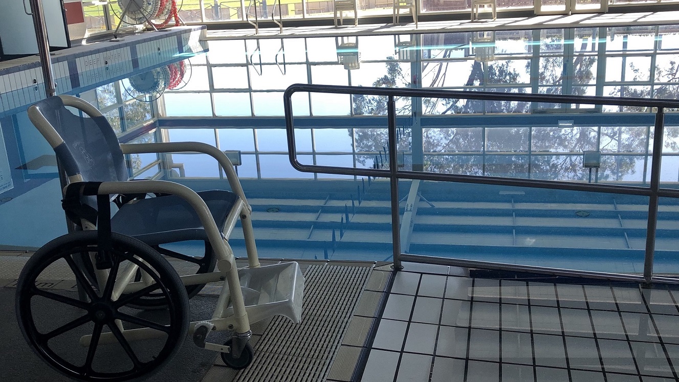 A closer look of the pool area where a blue and white aquatic wheelchair is placed on the side of the pool. The blue water shows the reflection of the white ceiling. The inner side of the pool’s coping has a text saying “Deep Water”. There are also silver pool stairs and an access ramp. A closer look of the pool area where a blue and white aquatic wheelchair is placed on the side of the pool. The blue water shows the reflection of the white ceiling. The inner side of the pool’s coping has a text saying “Deep Water”. There are also silver pool stairs and an access ramp.