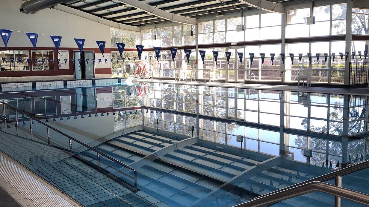 The view of the pool area where it’s showing the entire pool body. The blue water shows the reflection of the white ceiling and a long silver access ramp is at the side of the pool. There are long hanging banners made of white string and triangular blue pennants where “Zoggs“is printed. The glass wall of the pool area will let you see the bright and relaxing view from outside. The view of the pool area where it’s showing the entire pool body. The blue water shows the reflection of the white ceiling and a long silver access ramp is at the side of the pool. There are long hanging banners made of white string and triangular blue pennants where “Zoggs“is printed. The glass wall of the pool area will let you see the bright and relaxing view from outside.