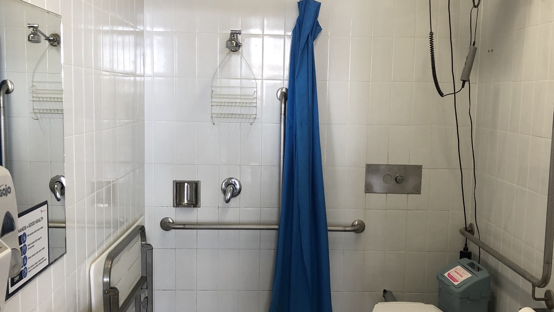 The leisure centre’s closer look of the white tiled restroom. It has silver grab handles at both sides, a silver built-in soap holder and a hanging accessory rack. A rectangular mirror is mounted on the left wall with a reminder posted on the bottom about the importance of washing hands. A blue folded curtain is hanging on the wall next to the shower head. A concealed cistern toilet suite is at the right corner where a trash bin is sitting next to it. The leisure centre’s closer look of the white tiled restroom. It has silver grab handles at both sides, a silver built-in soap holder and a hanging accessory rack. A rectangular mirror is mounted on the left wall with a reminder posted on the bottom about the importance of washing hands. A blue folded curtain is hanging on the wall next to the shower head. A concealed cistern toilet suite is at the right corner where a trash bin is sitting next to it.