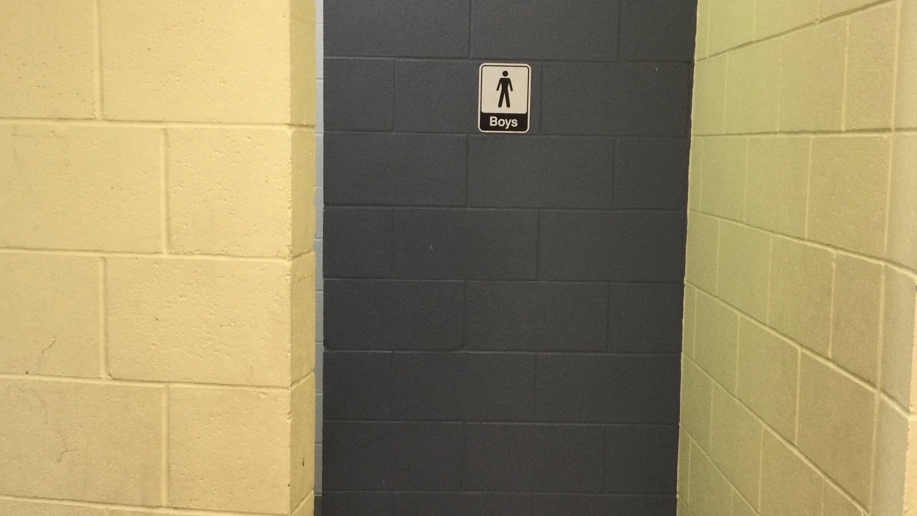 The male restroom's grey concrete wall has white and black signage with a male representation and the word “Boys”, while the yellow walls have a similar concrete design pattern. The male restroom's grey concrete wall has white and black signage with a male representation and the word “Boys”, while the yellow walls have a similar concrete design pattern.