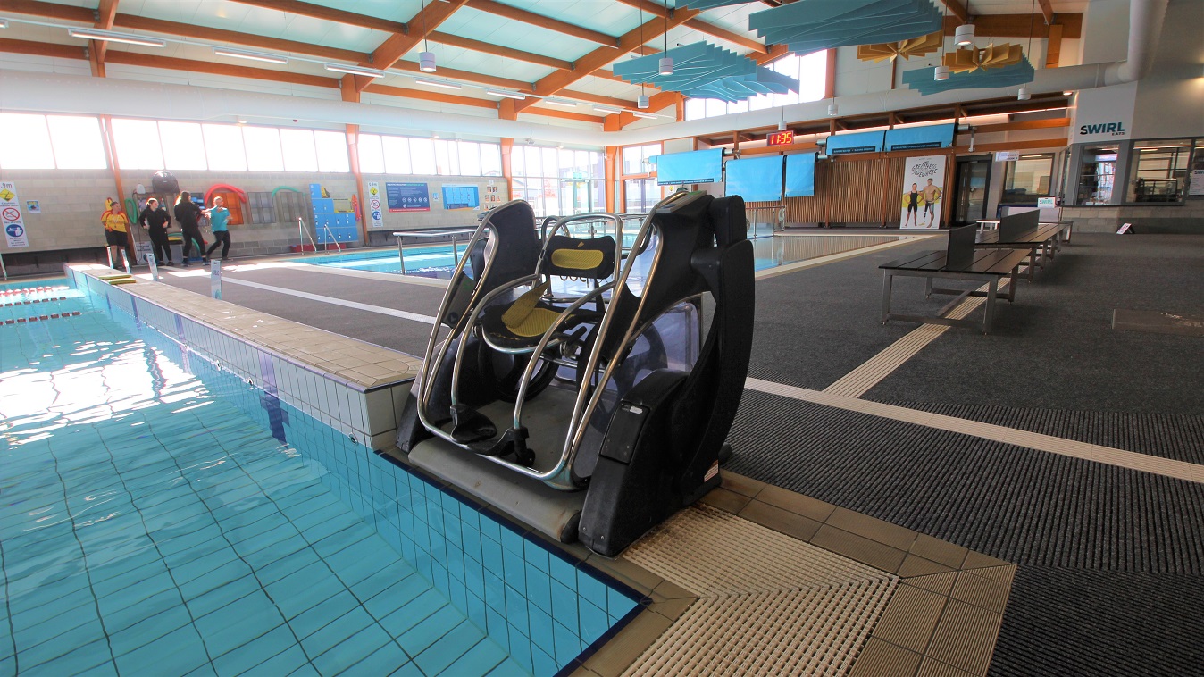 Aquatic wheelchair in the Aquatic facility at SWIRL. It is placed near the railed entrance ramp of a three-lane (6m x 12.5m) program pool that is 0.9m in depth. A digital clock can be seen placed on the upper wall of the facility. Separating the three-lane (6 m x 12.5m) program pool and the shallow pool found near the entrance door is a steel frame with a glass wall. Aquatic wheelchair in the Aquatic facility at SWIRL. It is placed near the railed entrance ramp of a three-lane (6m x 12.5m) program pool that is 0.9m in depth. A digital clock can be seen placed on the upper wall of the facility. Separating the three-lane (6 m x 12.5m) program pool and the shallow pool found near the entrance door is a steel frame with a glass wall.