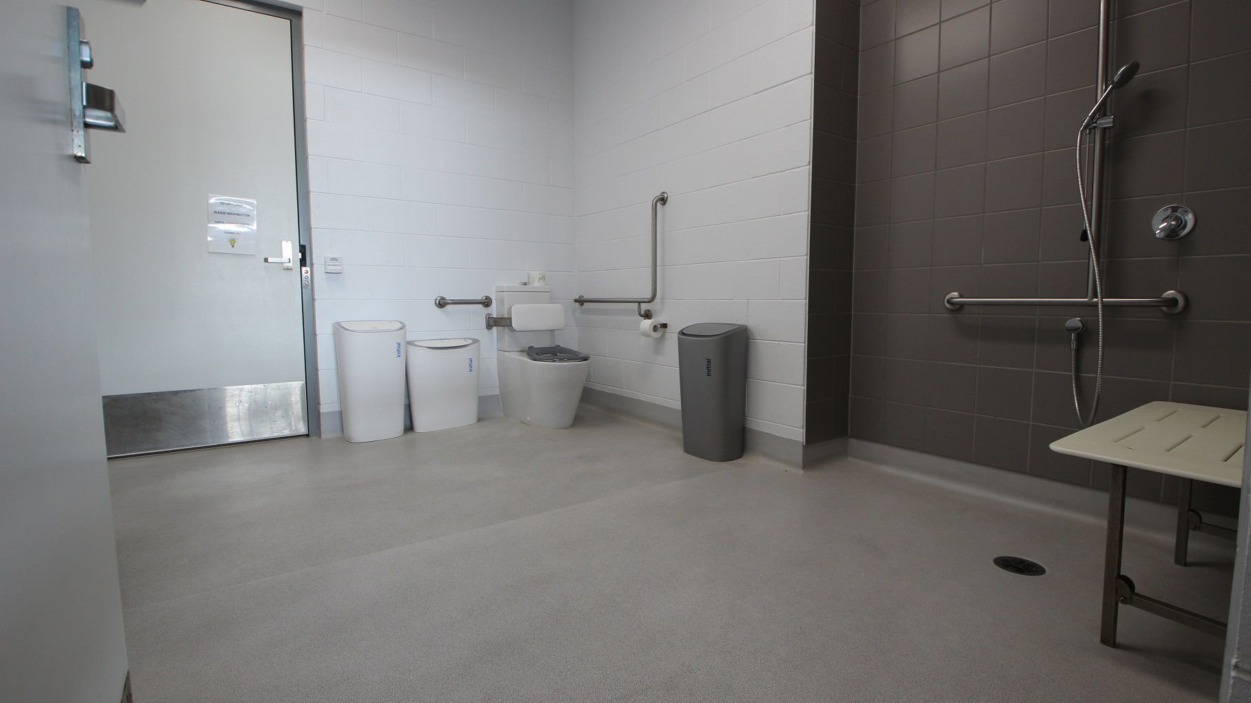 Inside look at the accessible change rooms at SWIRL. The shower area, equipped with grab bars and a handheld shower head, is painted grey. On the back is the toilet seat with a white cushion for the back. Mounted on the white tiled wall near it are grab bars and a toilet holder with a roll of toilet paper. On the focal point at the back is another door. Inside look at the accessible change rooms at SWIRL. The shower area, equipped with grab bars and a handheld shower head, is painted grey. On the back is the toilet seat with a white cushion for the back. Mounted on the white tiled wall near it are grab bars and a toilet holder with a roll of toilet paper. On the focal point at the back is another door.