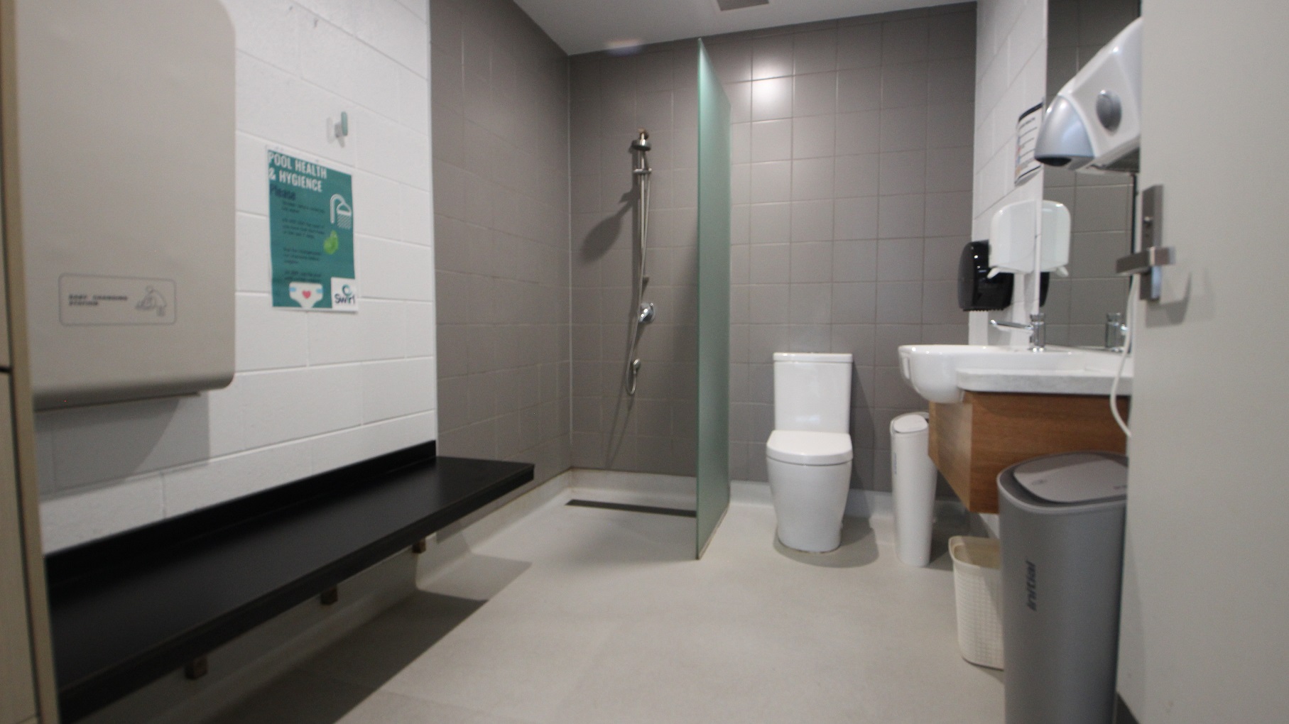 Inside the family change room at SWIRL. Mounted on the left white-painted brick wall is a black bench. The shower area and toilet seat are separated by a glass partition and the brick wall is painted in grey. On the right side are a wooden wash basin, a large mirror, tissue for hands, and a hand dryer Inside the family change room at SWIRL. Mounted on the left white-painted brick wall is a black bench. The shower area and toilet seat are separated by a glass partition and the brick wall is painted in grey. On the right side are a wooden wash basin, a large mirror, tissue for hands, and a hand dryer