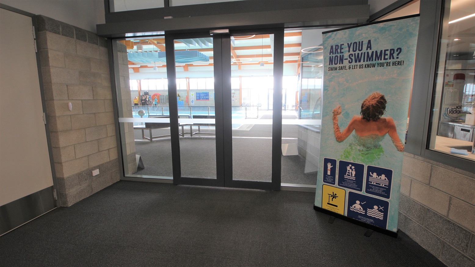 Automatic double glass entrance doors of the Aquatic facility inside SWIRL. The brick wall of this corridor has grey with a bit of dark portion at the bottom. On the right is a large poster with a picture of the child in a pool with the precautionary measures that need to be remembered by guests on the bottom. Automatic double glass entrance doors of the Aquatic facility inside SWIRL. The brick wall of this corridor has grey with a bit of dark portion at the bottom. On the right is a large poster with a picture of the child in a pool with the precautionary measures that need to be remembered by guests on the bottom.