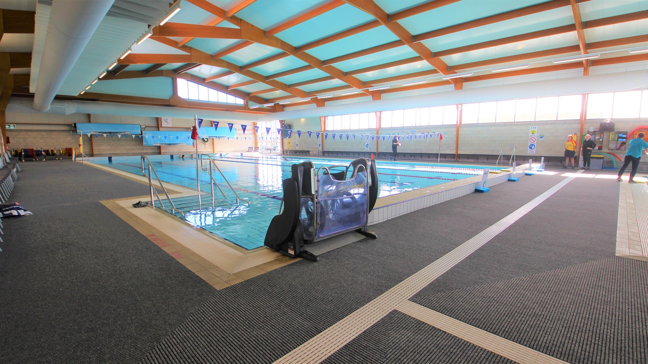 There is a PoolPod accessible hoist in the six-lane, 25-metre indoor pool that starts at 0.9m depth through to 1.8m depth with a 1.2m deep zone. The lanes are separated by red and blue floating ropes. It can be found next to the elevated section at the starting line of the pool and opposite the pool stairs with handrails. Intersecting horizontally to the pool is a string of hanging blue flags tied to opposite poles on the sides of the pool. There is a PoolPod accessible hoist in the six-lane, 25-metre indoor pool that starts at 0.9m depth through to 1.8m depth with a 1.2m deep zone. The lanes are separated by red and blue floating ropes. It can be found next to the elevated section at the starting line of the pool and opposite the pool stairs with handrails. Intersecting horizontally to the pool is a string of hanging blue flags tied to opposite poles on the sides of the pool.