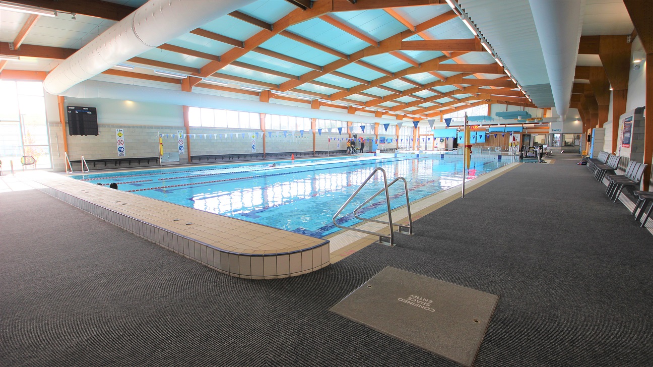 Opposite end of the 25-metre indoor pool that starts at 0.9m depth through to 1.8m depth with a 1.2m deep zone. The lanes are separated by red and blue floating ropes. There is a ladder found on both ends of the pool. This side of the pool has a string of hanging blue and white flags tied to opposite poles on the sides of the pool. Grey chairs can be seen on the right. Opposite end of the 25-metre indoor pool that starts at 0.9m depth through to 1.8m depth with a 1.2m deep zone. The lanes are separated by red and blue floating ropes. There is a ladder found on both ends of the pool. This side of the pool has a string of hanging blue and white flags tied to opposite poles on the sides of the pool. Grey chairs can be seen on the right.