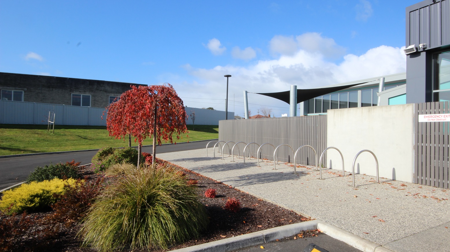 A number of inverted U-shape metal bike racks located on a rough surface of the cemented pathway near a garden allotment outside the grey fence and white wall. Inside the garden space are bushes of different types of green, yellow, green, brown, and red. Opposite the facility is a grey building with glass windows and grey, tall walls. A number of inverted U-shape metal bike racks located on a rough surface of the cemented pathway near a garden allotment outside the grey fence and white wall. Inside the garden space are bushes of different types of green, yellow, green, brown, and red. Opposite the facility is a grey building with glass windows and grey, tall walls.