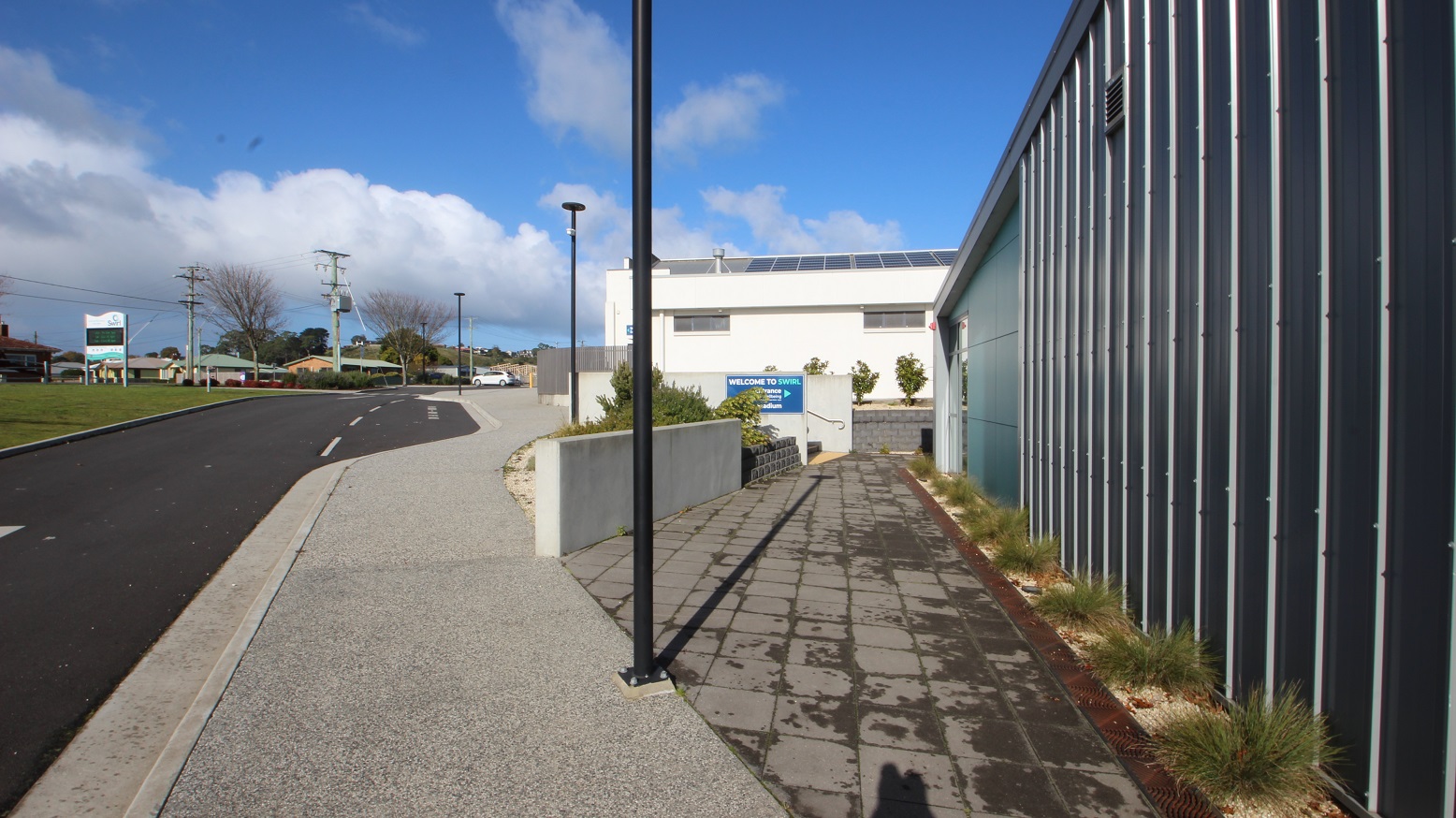 Crossing the zebra lanes and turning right off the smooth cemented pathway with a black lamp post is a grey tile floor to the SWIRL entrance. Tall grasses are linearly arranged against the light green and black segmented walls of the facility. Heading straight from this pathway and to the left is a Welcome to SWIRL sign placed on the short white wall next to a staircase with metal handrails. Crossing the zebra lanes and turning right off the smooth cemented pathway with a black lamp post is a grey tile floor to the SWIRL entrance. Tall grasses are linearly arranged against the light green and black segmented walls of the facility. Heading straight from this pathway and to the left is a Welcome to SWIRL sign placed on the short white wall next to a staircase with metal handrails.