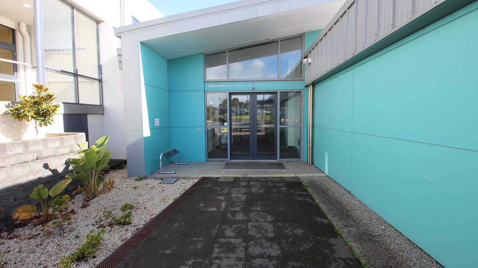 Grey tiled pathway leading to the entrance automatic, double glass doors of SWIRL. There is a type of plant from the banana family due to its leaves in the garden box. Prohibited things inside the facility are displayed on the right glass door (no smoking sign, no pets allowed, no running, etc). On the top of the glass doors is SMITHTON WELLBEING INDOOR RECREATION & LEISURE. Found on the wall at the end of the entrance ramp of the white building is an Authorised Entry Only sign. Grey tiled pathway leading to the entrance automatic, double glass doors of SWIRL. There is a type of plant from the banana family due to its leaves in the garden box. Prohibited things inside the facility are displayed on the right glass door (no smoking sign, no pets allowed, no running, etc). On the top of the glass doors is SMITHTON WELLBEING INDOOR RECREATION & LEISURE. Found on the wall at the end of the entrance ramp of the white building is an Authorised Entry Only sign.