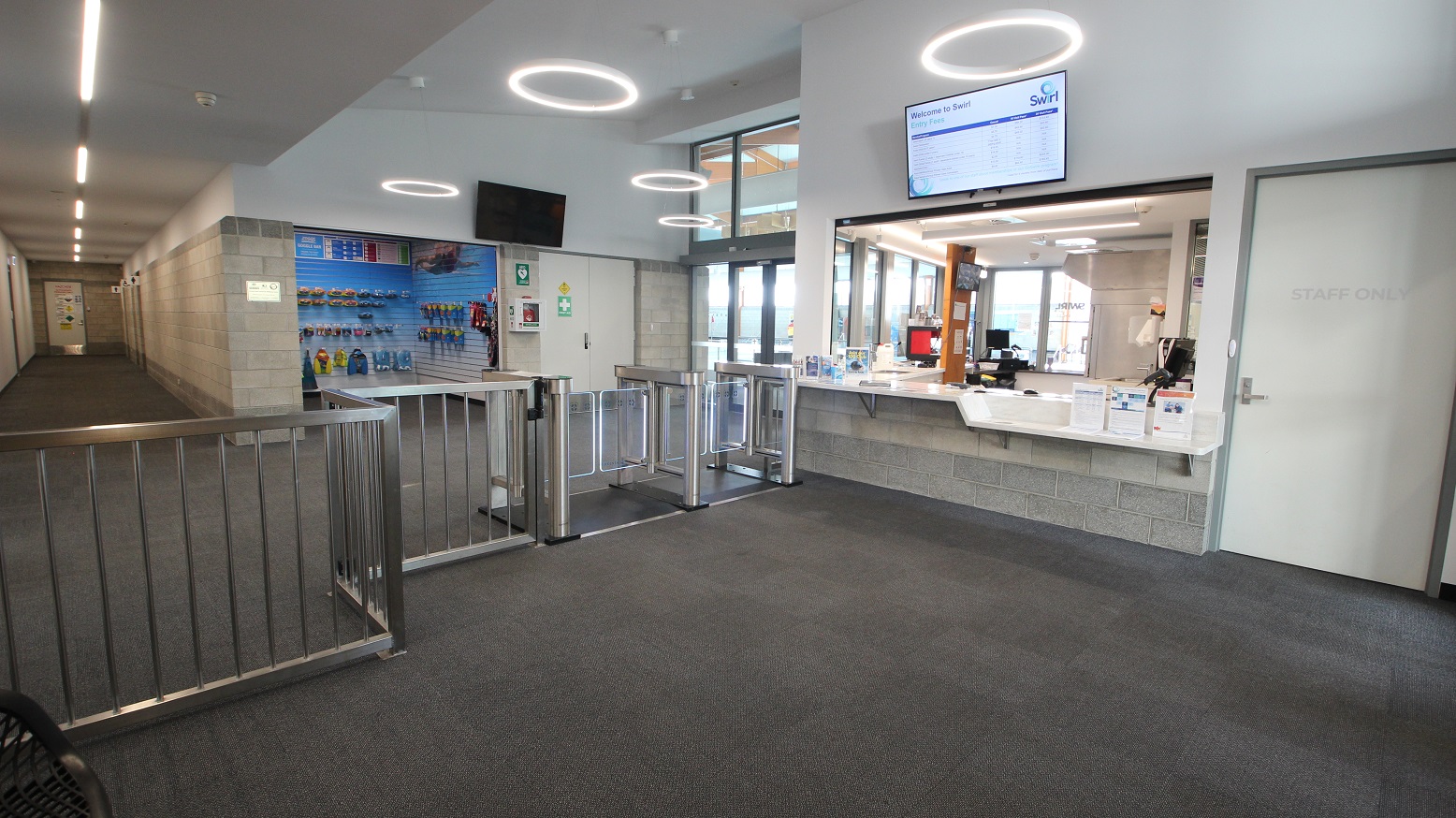 Turnstiles, including an accessible turnstile that leads to a small retail space selling a range of aquatic gear. Staff can go to the reception area through a white door with a STAFF ONLY sign. On the upper white wall of the reception desk is a television showing information about SWIRL. The retail space has a grey brick wall. There is a corridor found on the left, next to the retail space. Turnstiles, including an accessible turnstile that leads to a small retail space selling a range of aquatic gear. Staff can go to the reception area through a white door with a STAFF ONLY sign. On the upper white wall of the reception desk is a television showing information about SWIRL. The retail space has a grey brick wall. There is a corridor found on the left, next to the retail space.
