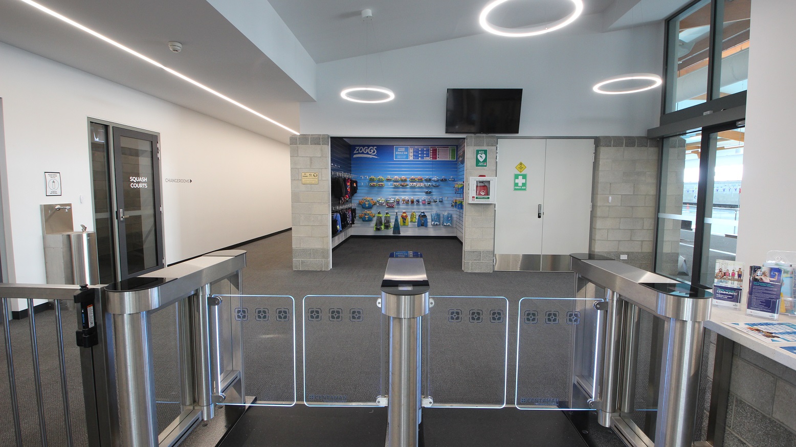 A closer look at the turnstiles, including an accessible turnstile that leads to a small retail space selling a range of aquatic gear. On the upper wall is a television. There is a white double door with a green first aid sign on the right of the retail space. There is a water fountain station accessible on the left corridor that leads to Changerooms. Right before the water fountain stain is the tinted door of SQUASH Courts. A closer look at the turnstiles, including an accessible turnstile that leads to a small retail space selling a range of aquatic gear. On the upper wall is a television. There is a white double door with a green first aid sign on the right of the retail space. There is a water fountain station accessible on the left corridor that leads to Changerooms. Right before the water fountain stain is the tinted door of SQUASH Courts.