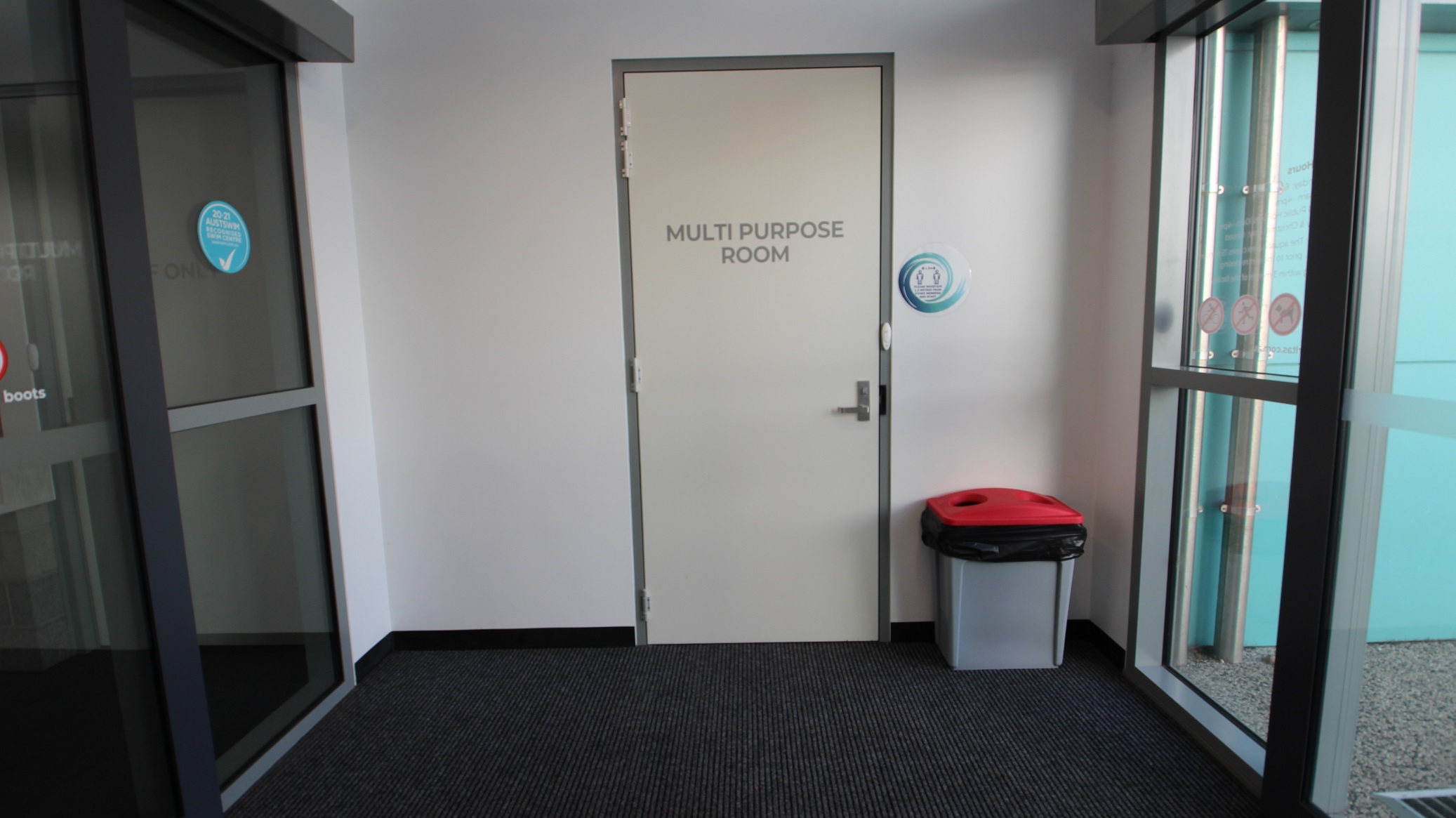 A small room with a grey carpeted floor and white walls. In the centre, a door labelled "MULTI PURPOSE ROOM" is visible. To the right of the door, a grey bin with a red lid is placed against the wall. On the left, there is a glass door with a sticker, and on the right, a large window with a view of a light blue exterior wall. The ceiling is not visible.