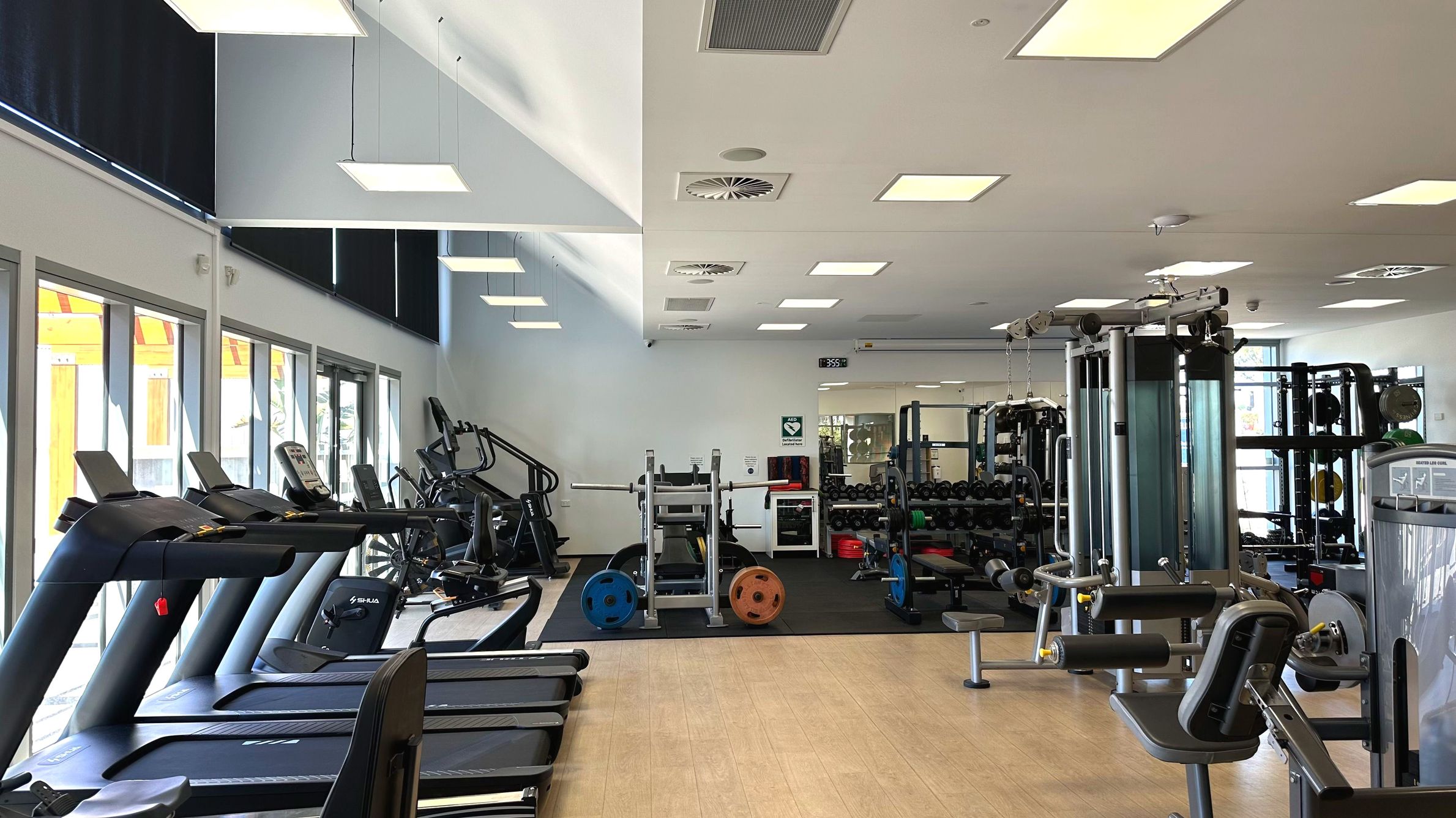A modern gym interior with a variety of exercise equipment. On the left, several treadmills are lined up facing large windows with black blinds. In the centre, weightlifting equipment, including a squat rack with colourful weight plates, is visible. To the right, there are various strength machines. The ceiling features recessed lighting and air vents. The floor transitions from light wood to black rubber matting in the weight area.