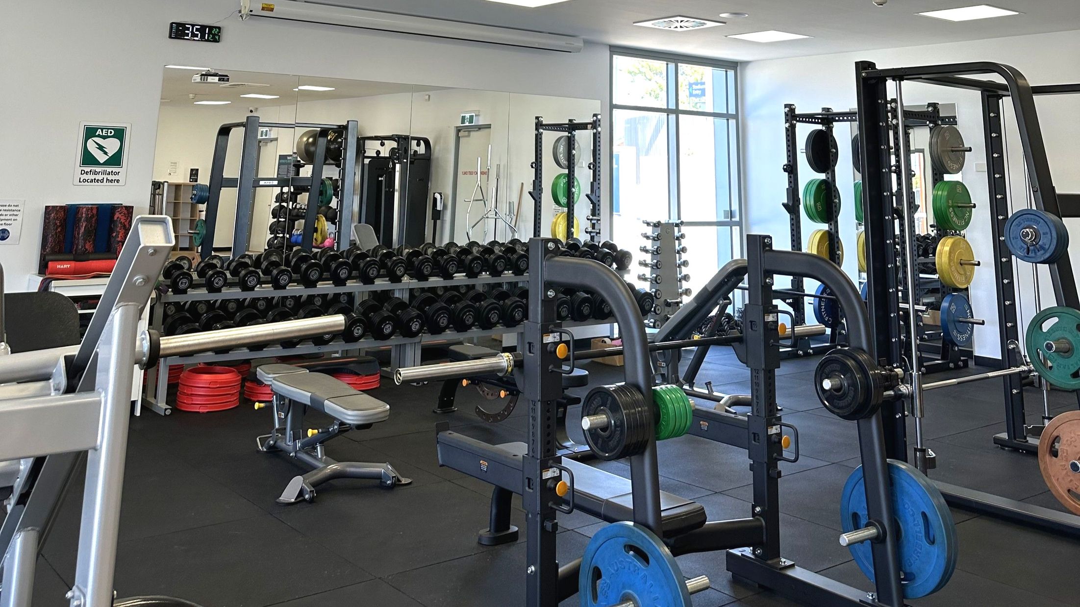 A gym room with various exercise equipment including weight racks, benches, and a variety of weights. A large mirror covers one wall, reflecting the equipment and adding depth to the space. A sign indicating a defibrillator is mounted on the wall. The floor is covered with black rubber mats, and a large window allows natural light to enter. The room appears organised and well-equipped for weight training.