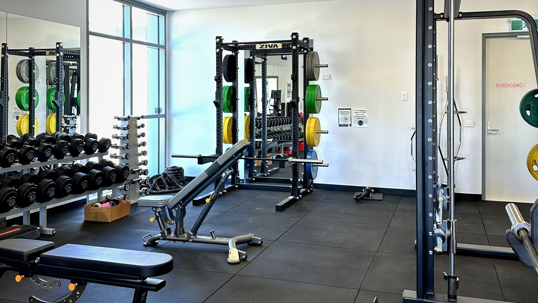 A gym room with various exercise equipment. On the left, there are racks of dumbbells and a vertical stand with weight plates in green, yellow, and black. A bench press is positioned in the centre, and a squat rack with a barbell is visible. The floor is covered with black rubber mats. A large window on the left wall allows natural light in. An emergency exit door is on the right wall, marked with a sign.