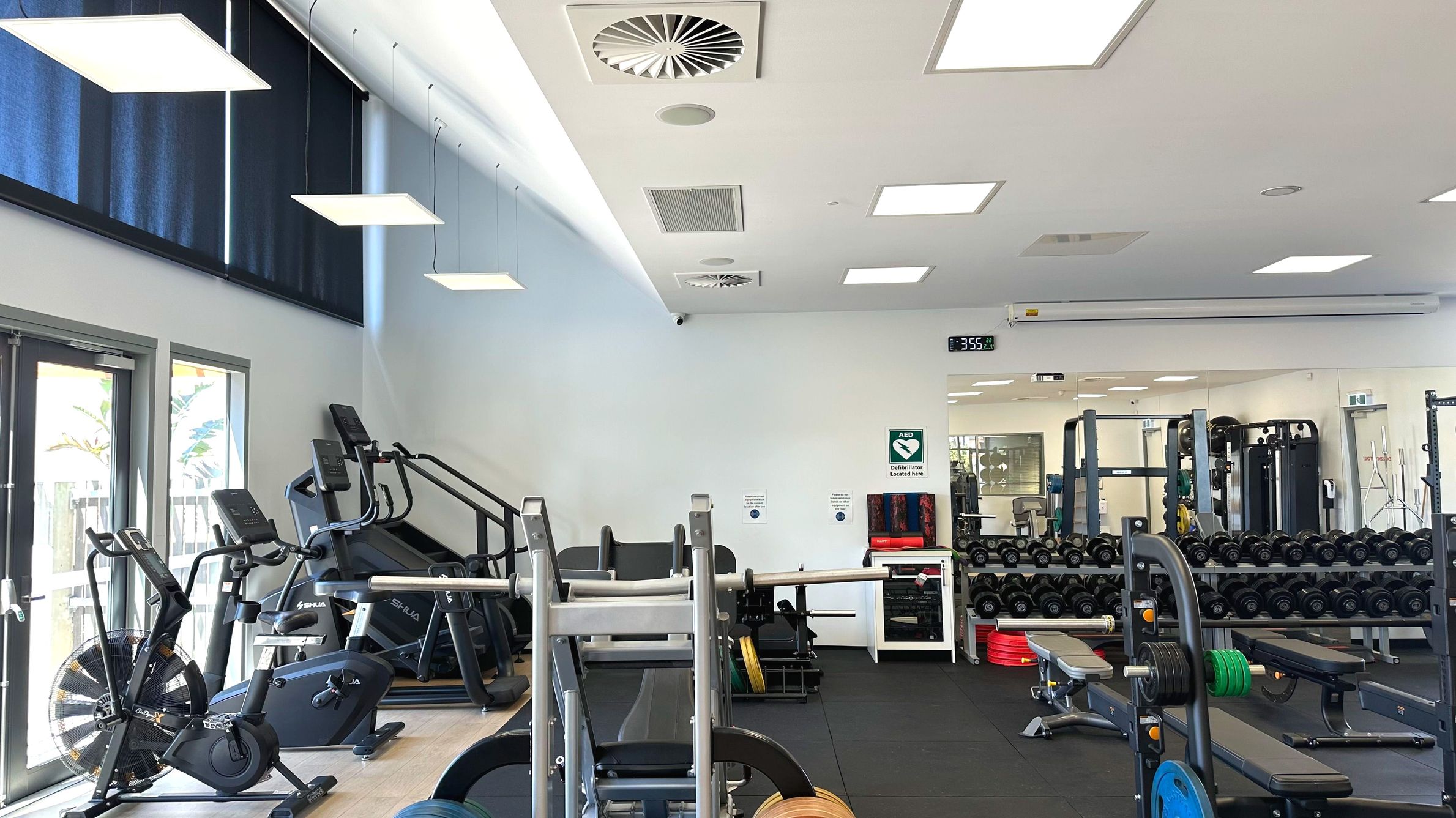 A gym interior with various exercise equipment including stationary bikes, treadmills, and weightlifting benches. The room features a mirrored wall reflecting the equipment, a digital clock displaying 3:55, and a sign indicating an AED location. The ceiling has recessed lighting and ventilation panels. The floor transitions from light wood to dark rubber mats. Large windows with blue blinds allow natural light in.