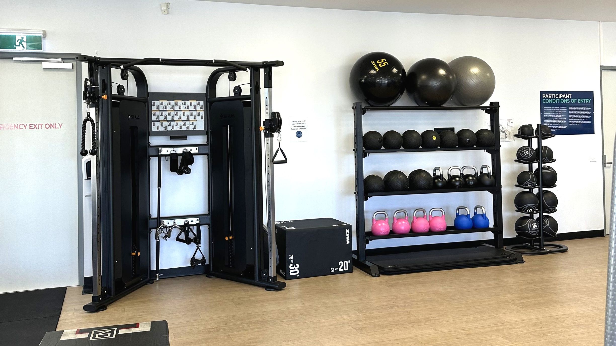 A gym area with a variety of exercise equipment. On the left, a multifunctional cable machine with various attachments is positioned next to a door marked 'Emergency Exit Only'. To the right, shelves hold large exercise balls, kettlebells in pink and blue, and medicine balls. A sign titled 'Participant Conditions of Entry' is visible on the wall. The floor is light wood, and the space feels open with white walls.