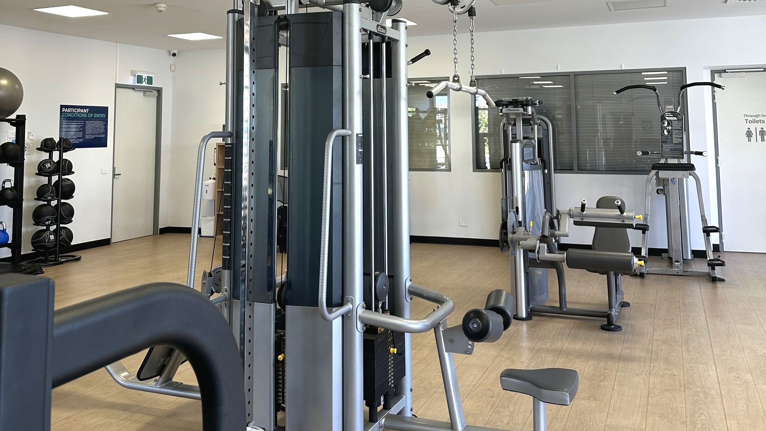 A gym room with various exercise equipment including a multi-station machine in the centre, a leg press, and a pull-up station. To the left, a rack holds medicine balls. The floor is light wood, and the walls are white with a door marked 'Toilets' on the right. A sign with 'Participant Conditions of Entry' is visible near another door. Large windows with blinds are on the back wall, allowing natural light.