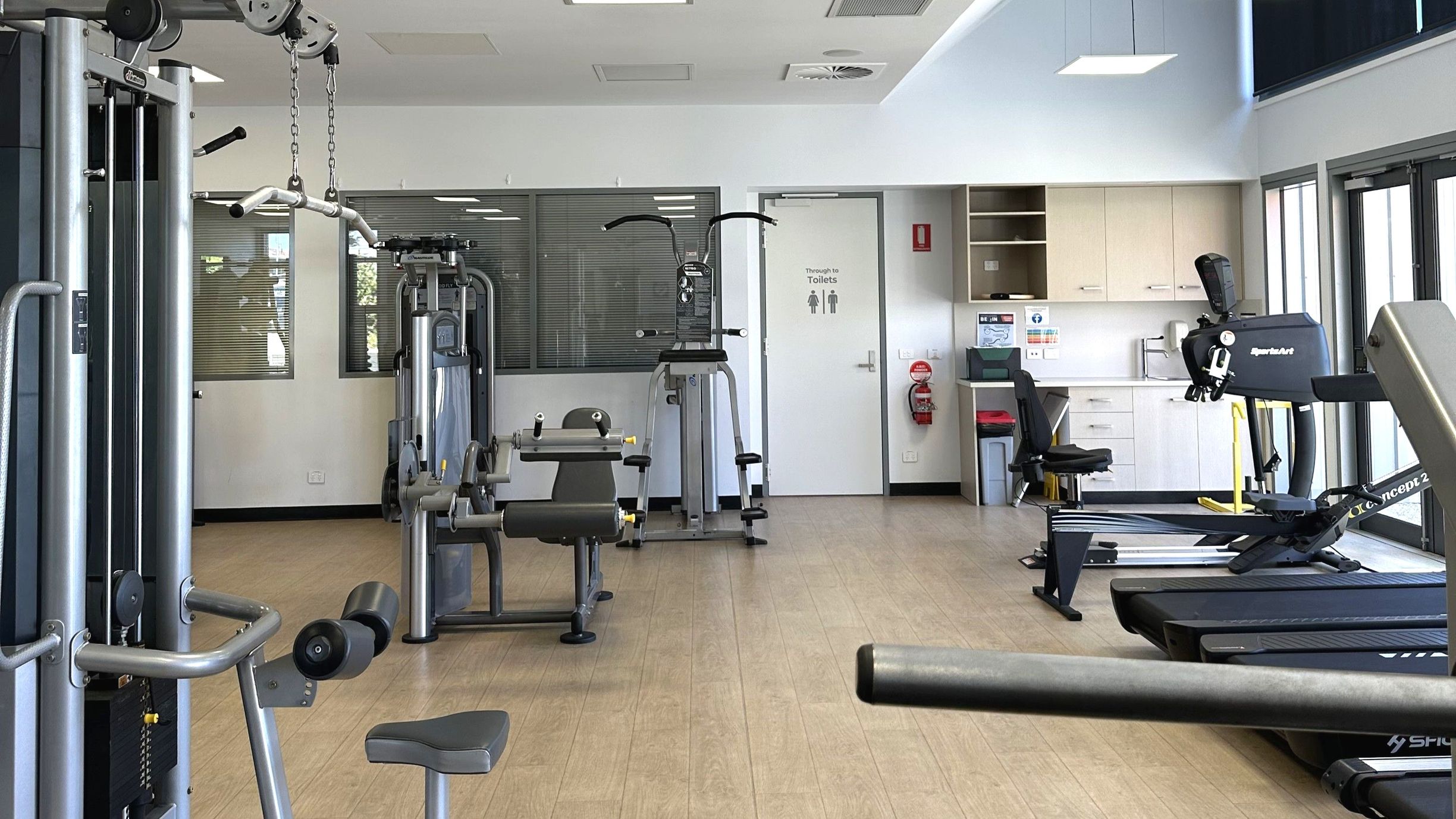 A gym room with various exercise equipment including a cable machine, a rowing machine, and a leg press. The floor is light wood, and the walls are white with large windows on the right. A door marked 'Through to Toilets' is visible at the back, next to a fire extinguisher. Cabinets and a sink are on the right wall. The space is well-lit with ceiling lights and natural light from the windows.