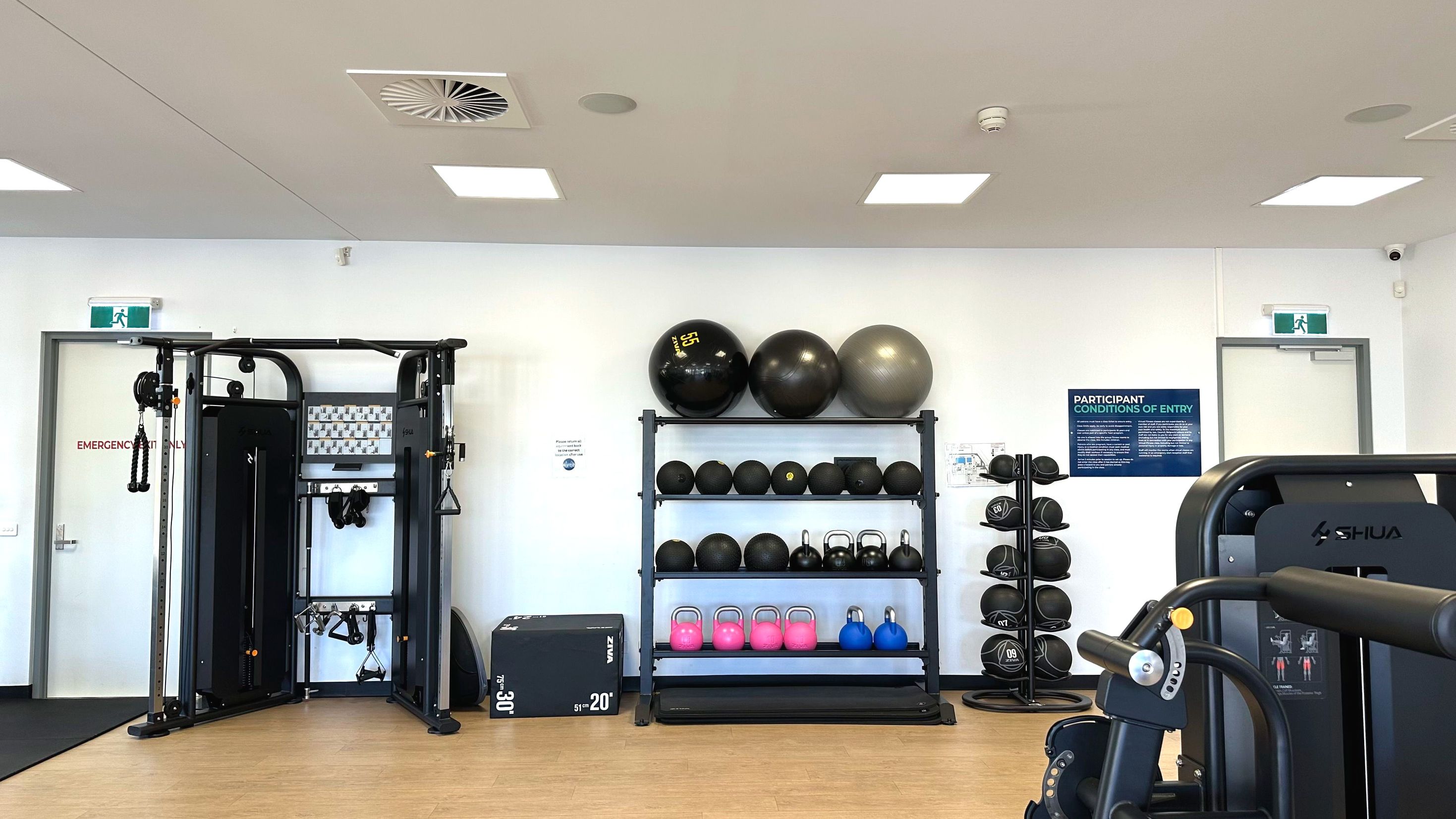 A gym room with a white wall featuring a variety of exercise equipment. On the left, a multi-functional cable machine stands next to a door marked 'Emergency Exit Only'. In the centre, a rack holds exercise balls, kettlebells, and medicine balls. To the right, a blue sign with text is visible above a stack of medicine balls. The floor is light wood, and the ceiling has recessed lighting and ventilation panels.