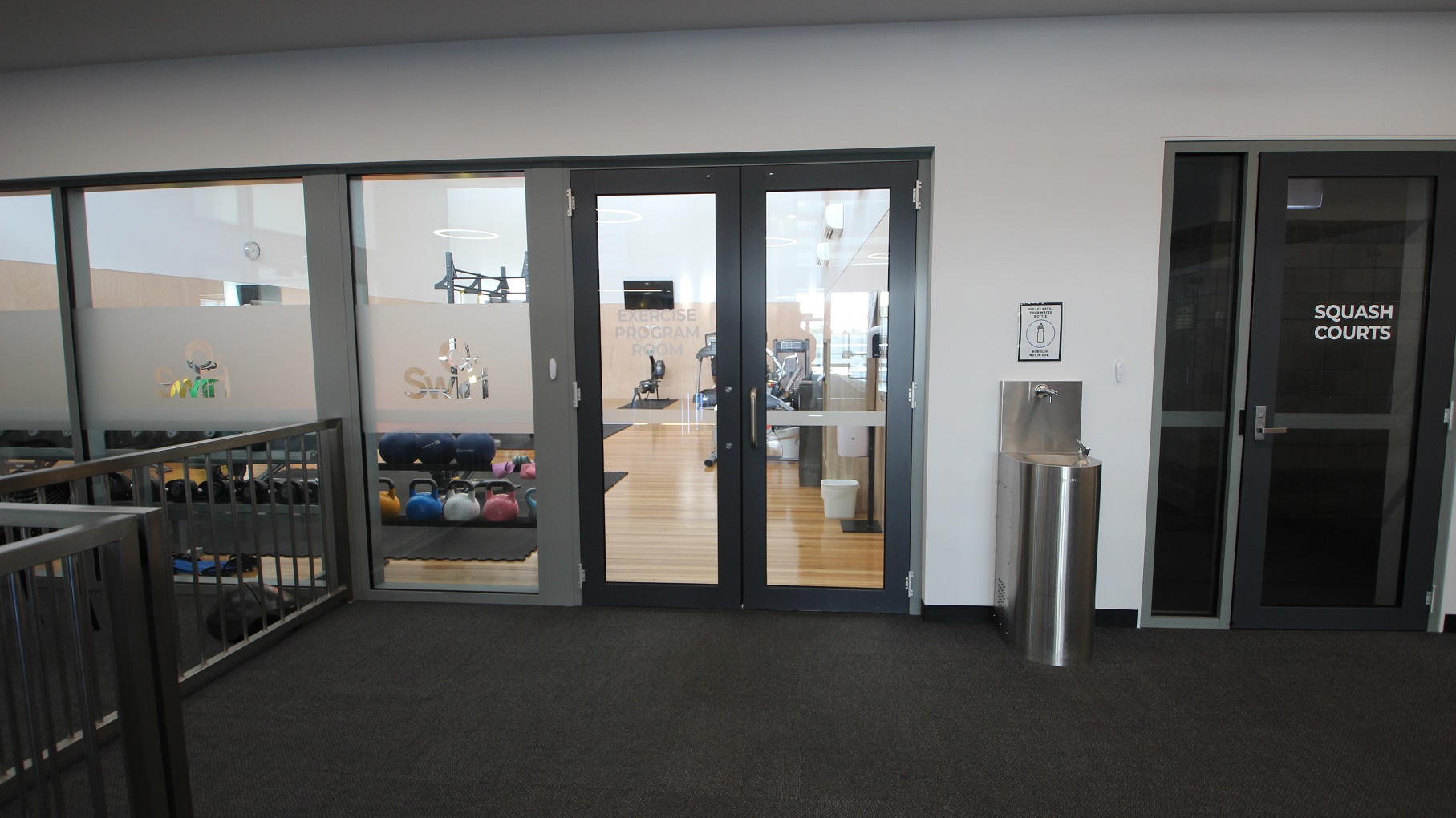 A multipurpose group exercise area. On the right, a door marked "Squash Courts" is visible. A water fountain and a bin are positioned between the doors. The floor is carpeted in dark grey, and the walls are white.