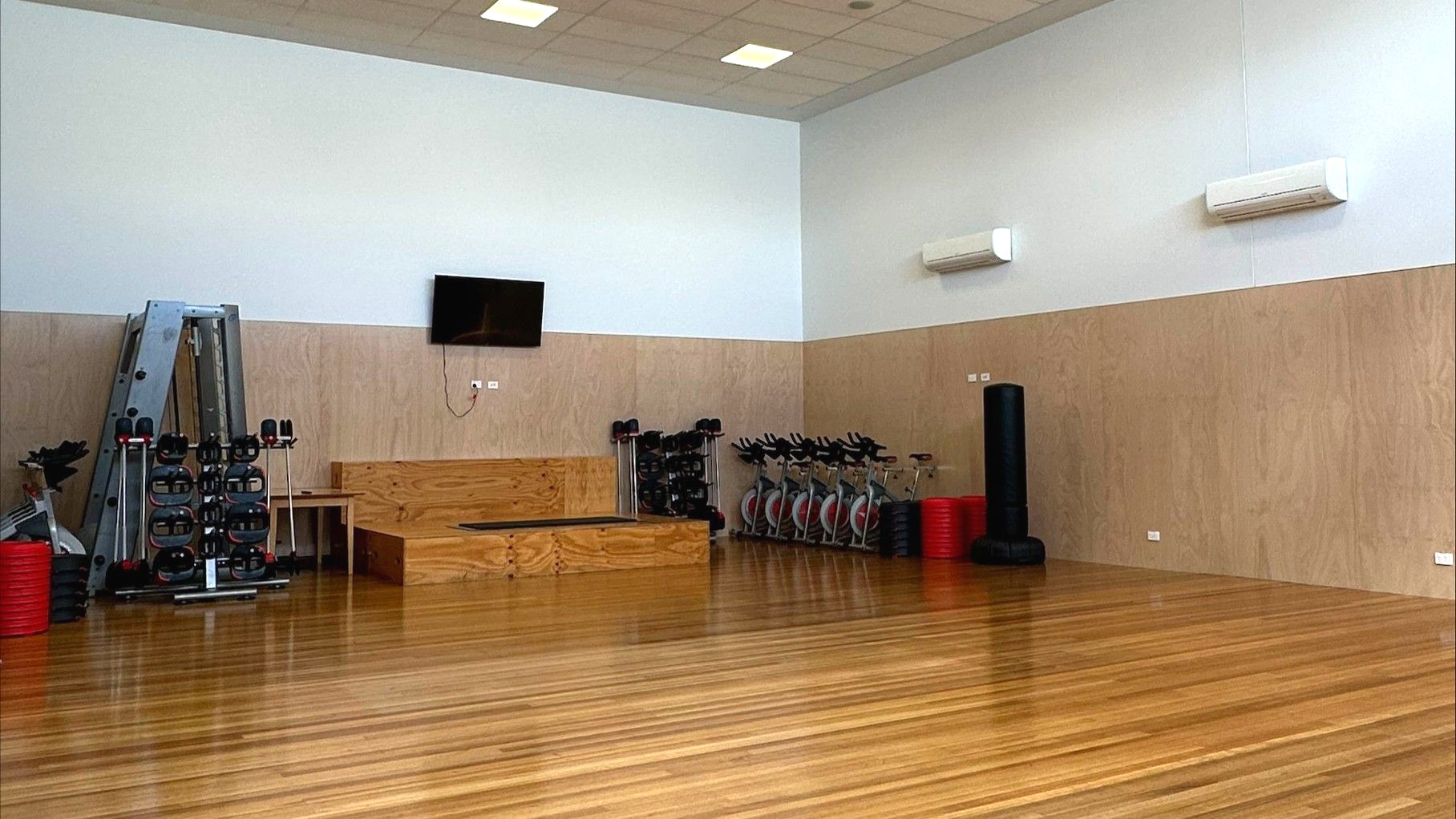 A spacious gym room with wooden flooring and light-coloured walls. On the left, there is a rack of dumbbells and a cable machine. In the centre, a wooden platform is positioned against the wall, with a wall-mounted TV above it. To the right, several stationary bikes are lined up, and a tall black punching bag stands nearby. The ceiling has recessed lighting, and two air conditioning units are mounted on the wall.