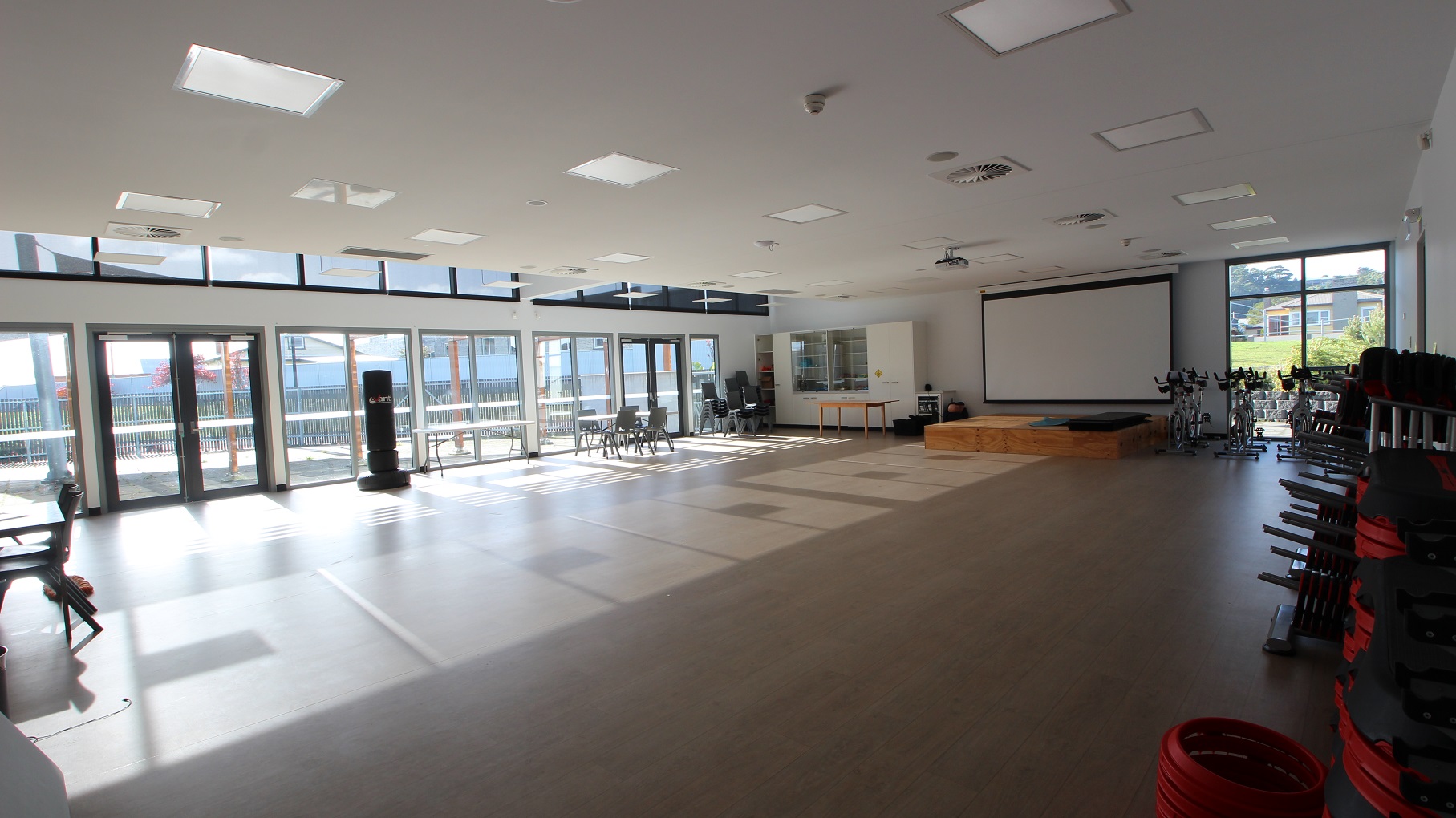 Inside the Multipurpose room is a large, open space. There is a wooden block in front of the large projector screen. On the left is a white cabinet with a stack of black chairs with steel legs. On the right are stationary bicycles for exercise. Natural light is filtered through the glass walls and windows. Inside the Multipurpose room is a large, open space. There is a wooden block in front of the large projector screen. On the left is a white cabinet with a stack of black chairs with steel legs. On the right are stationary bicycles for exercise. Natural light is filtered through the glass walls and windows.