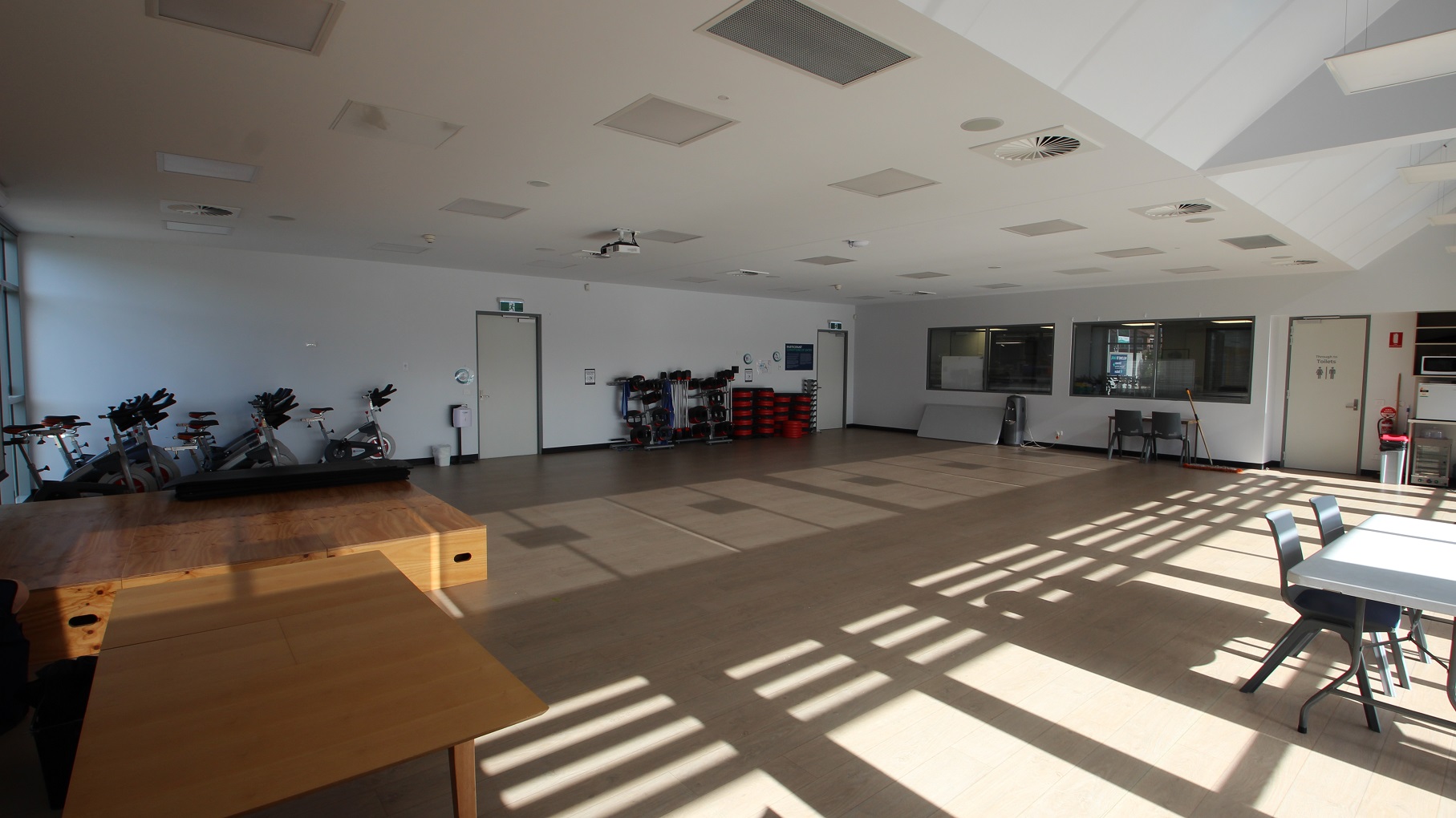 There are three doors inside the Multipurpose room. There is a wooden block in front of the large projector screen, and white tables with grey chairs are accessible in the room. On the right are stationary bicycles for exercise. Natural light is filtered through the glass walls and windows. There are three doors inside the Multipurpose room. There is a wooden block in front of the large projector screen, and white tables with grey chairs are accessible in the room. On the right are stationary bicycles for exercise. Natural light is filtered through the glass walls and windows.