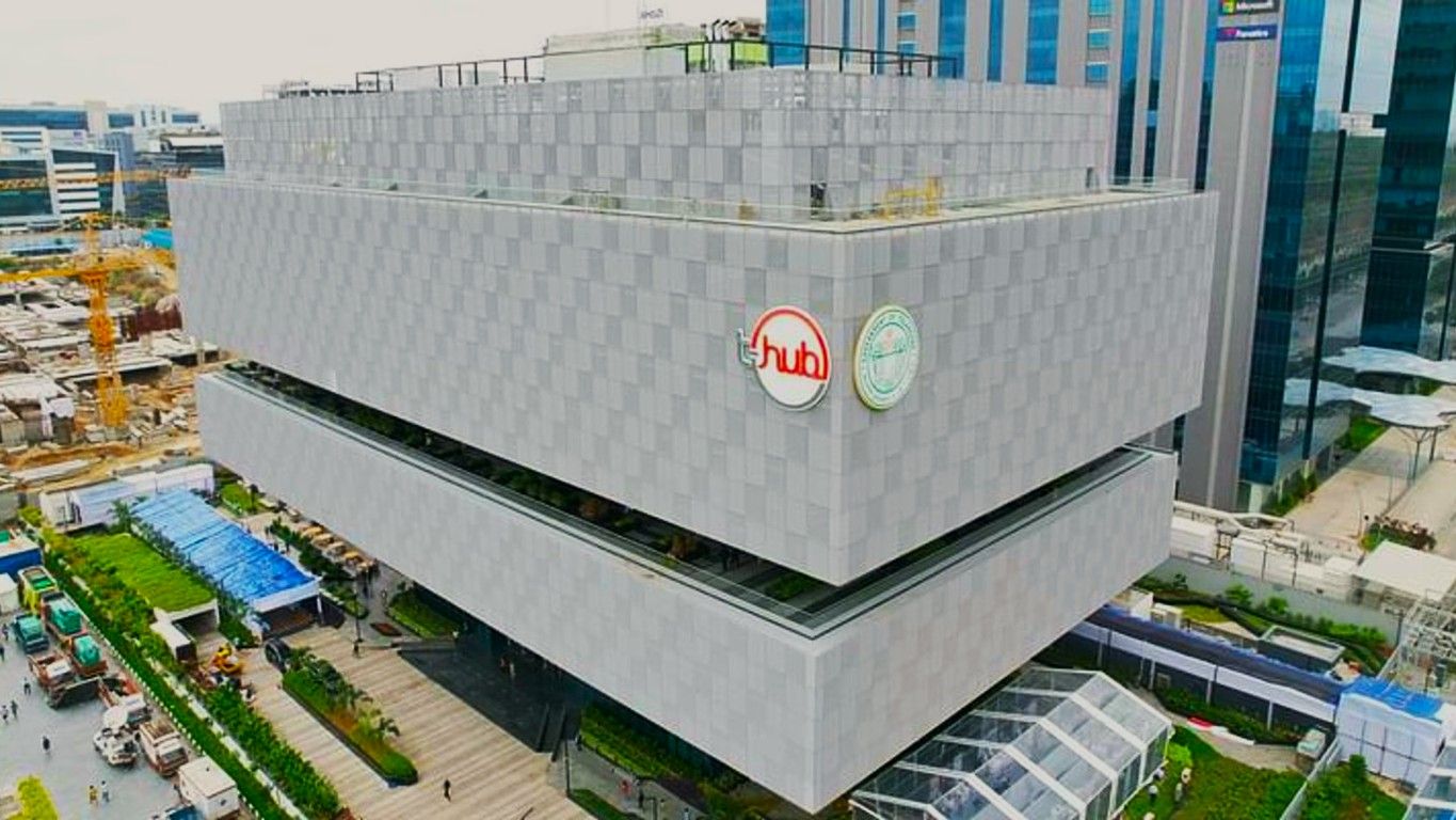 A modern, multi-storey building with a grey, checkered facade and two circular logos on the side. The structure has a unique, staggered design with a green terrace visible on one level. Surrounding the building are other high-rise structures with glass exteriors. In the foreground, there is a landscaped area with pathways, greenery, and a few vehicles parked. Construction activity is visible to the left, with cranes and building materials.