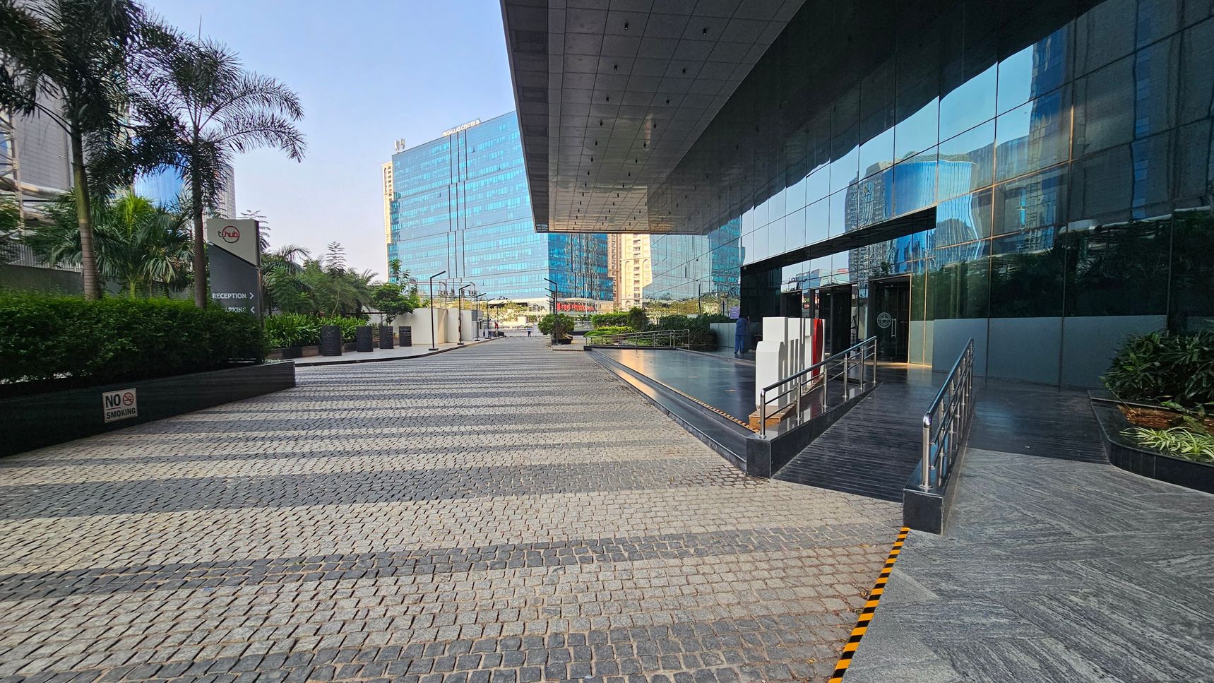 A modern urban setting with a cobblestone pathway leading to a building with reflective glass walls on the right. The building has a covered entrance with a ramp and steps. On the left, there are palm trees and a sign with directional arrows. In the background, tall glass buildings are visible. The area is landscaped with greenery, and a "No Smoking" sign is present. The ceiling of the entrance is sleek and metallic.